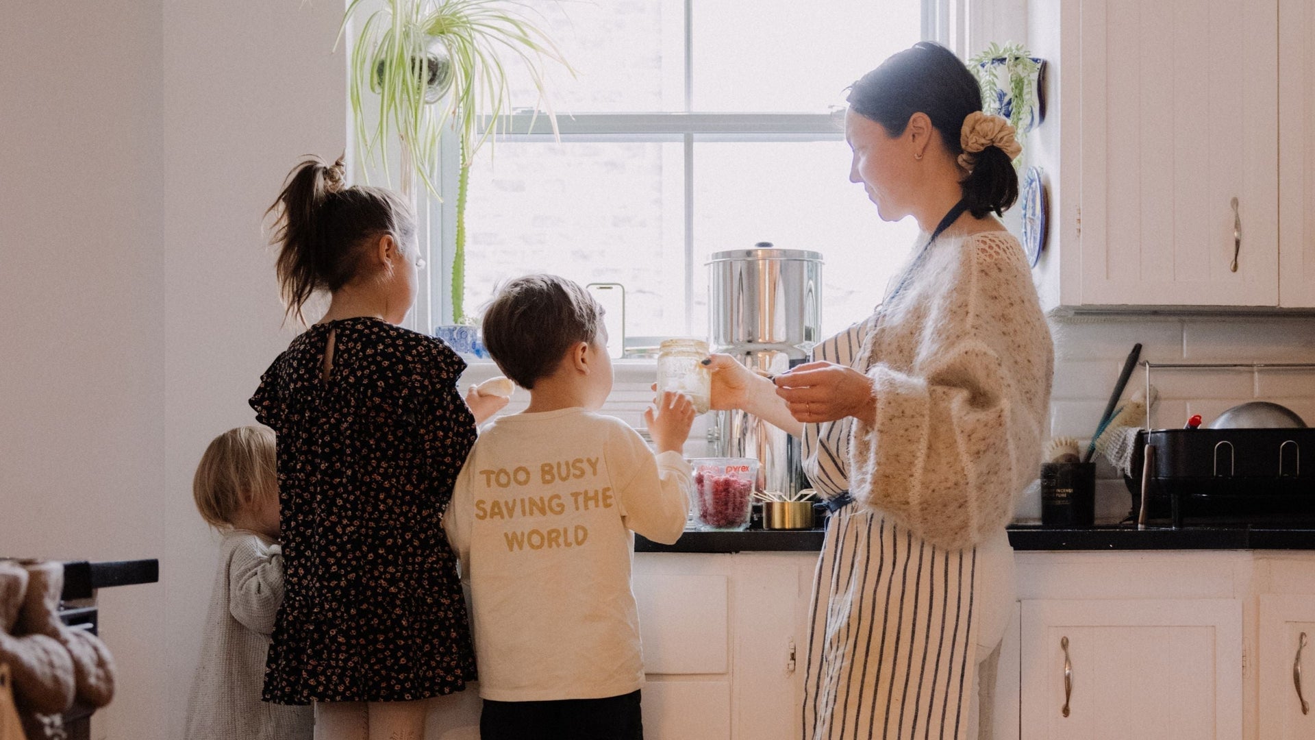 A woman and three young children preparing food together in a bright kitchen. One child wears a shirt with the words 'Too busy saving the world' while the woman holds a jar of ingredients near a sunny window.