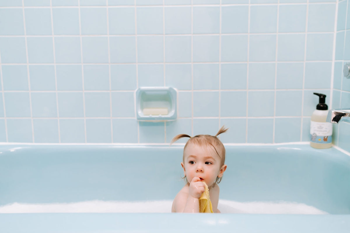 A toddler with small pigtails sitting in a blue bubble bath, holding a yellow washcloth up to their mouth, with a bottle of The Unscented Company Gentle Baby Wash & Shampoo sitting on the edge of the tub.