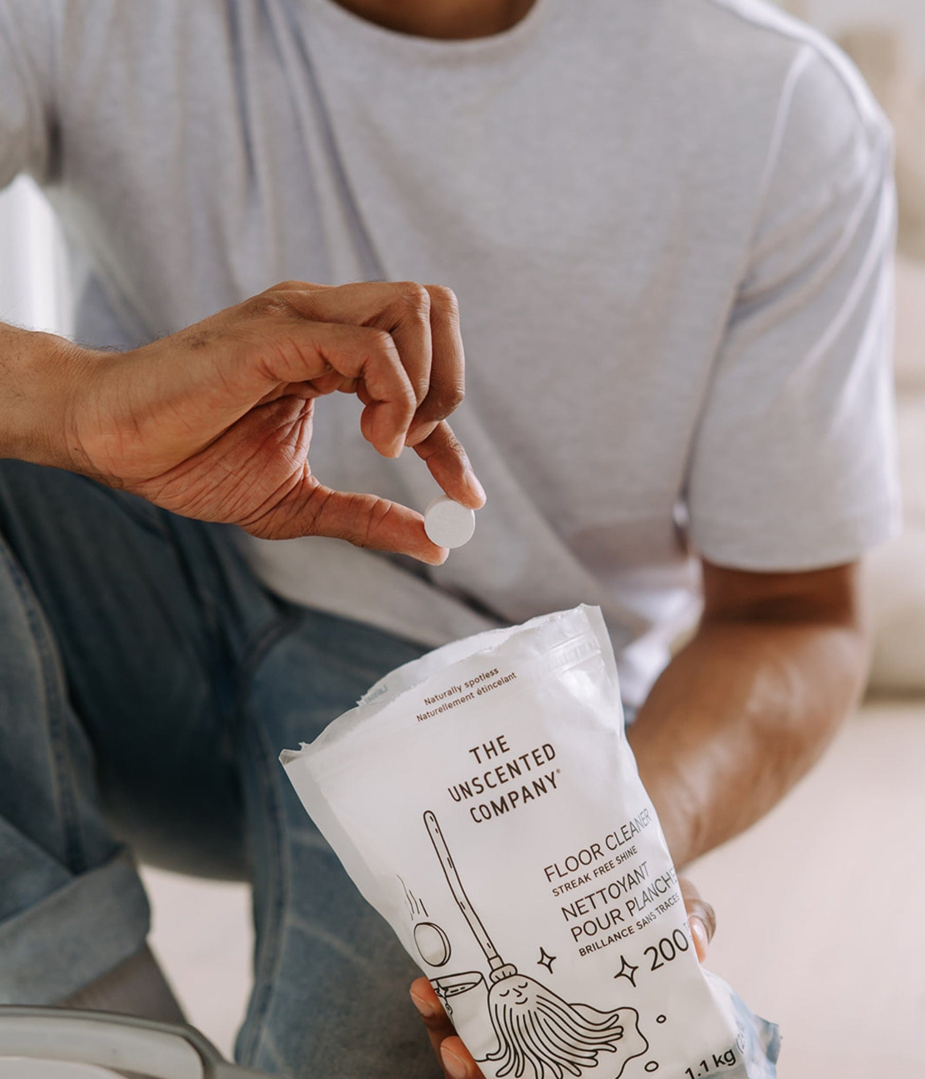 Gros plan sur la main d’un homme tenant une pastille nettoyante pour plancher au-dessus d’un sachet ouvert de nettoyant pour plancher de The Unscented Company. Le sachet est blanc avec des illustrations et une étiquette bilingue.