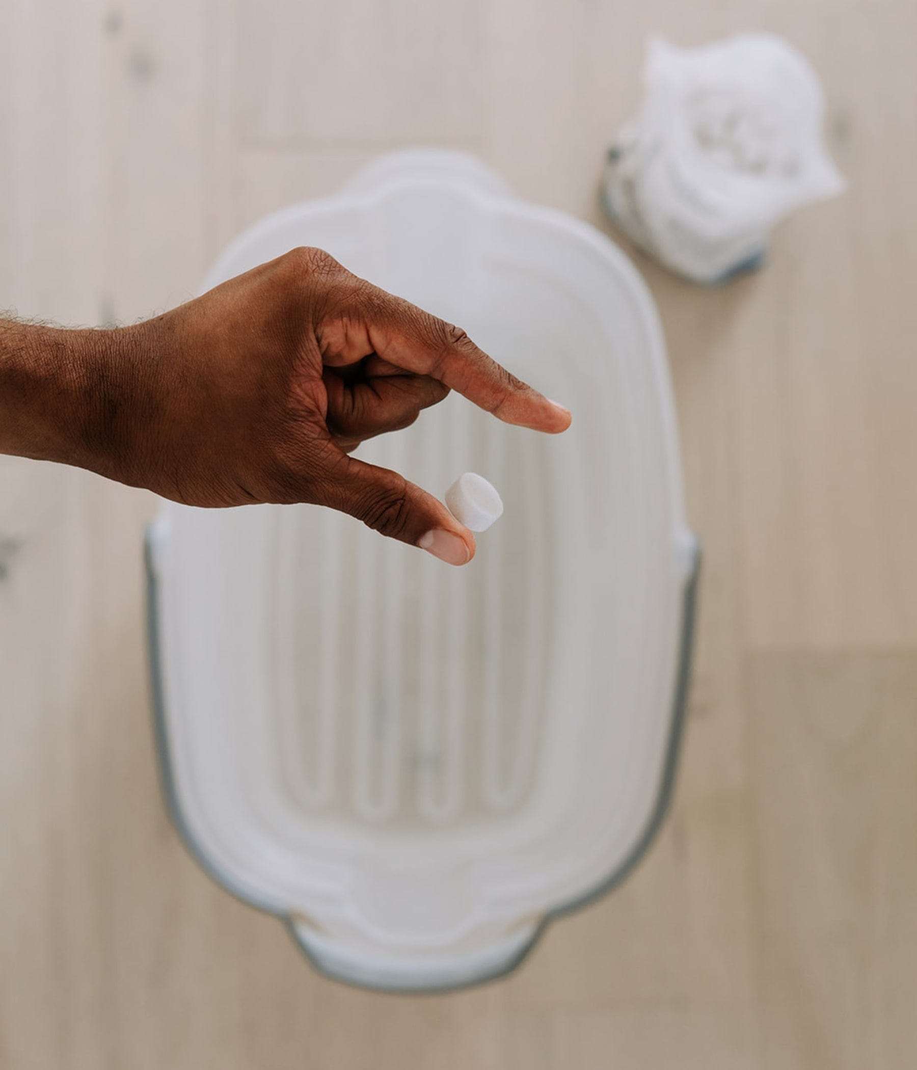 Une main tient une pastille nettoyante blanche au-dessus d’un seau rempli d’eau. En arrière-plan, un sachet de nettoyant pour plancher The Unscented Company est légèrement flou, posé sur un plancher de bois pâle.