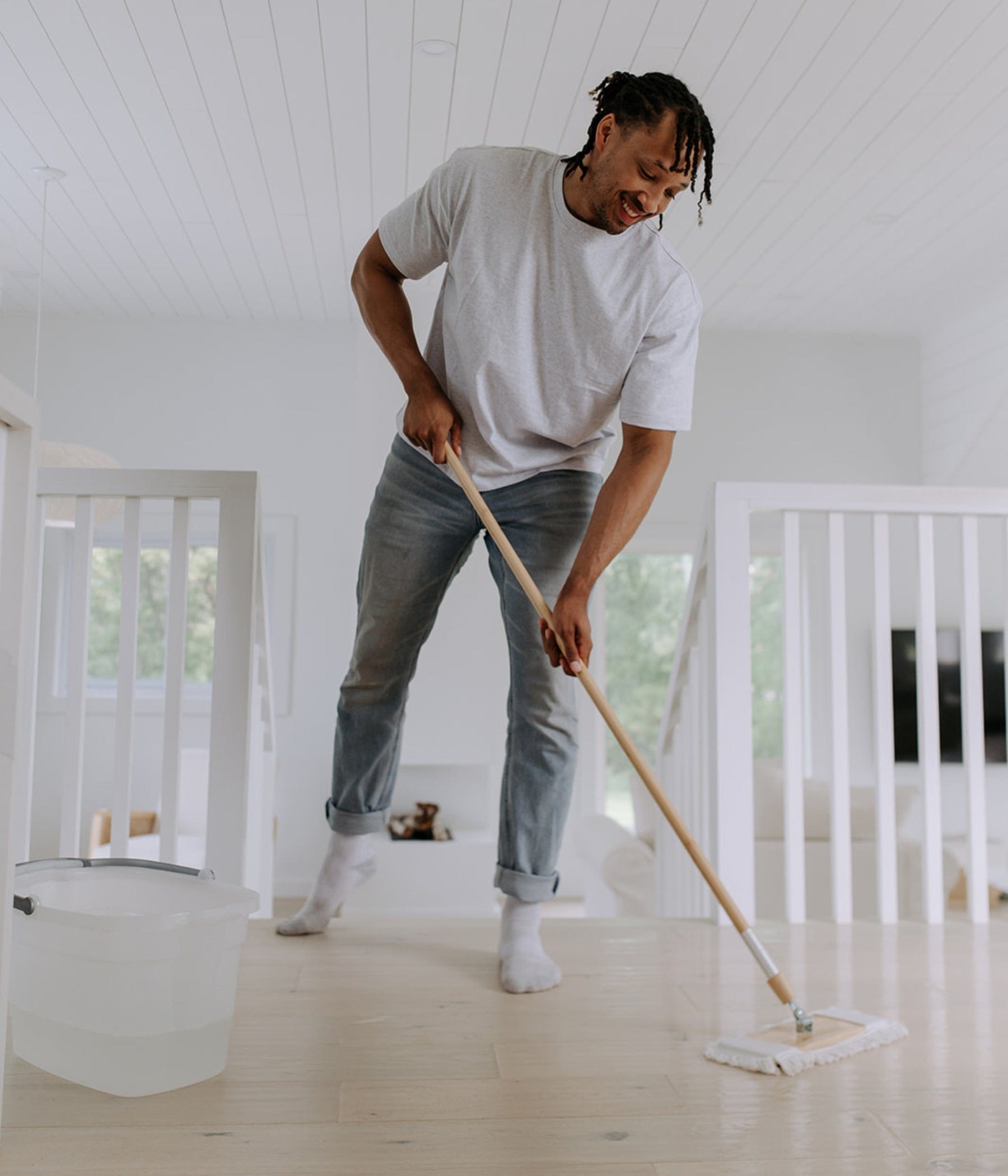 Un homme souriant passe la vadrouille sur un plancher de bois dans une maison lumineuse et minimaliste. Un seau blanc rempli d’eau est posé à proximité. L’environnement est épuré, aux murs et rampes blancs.