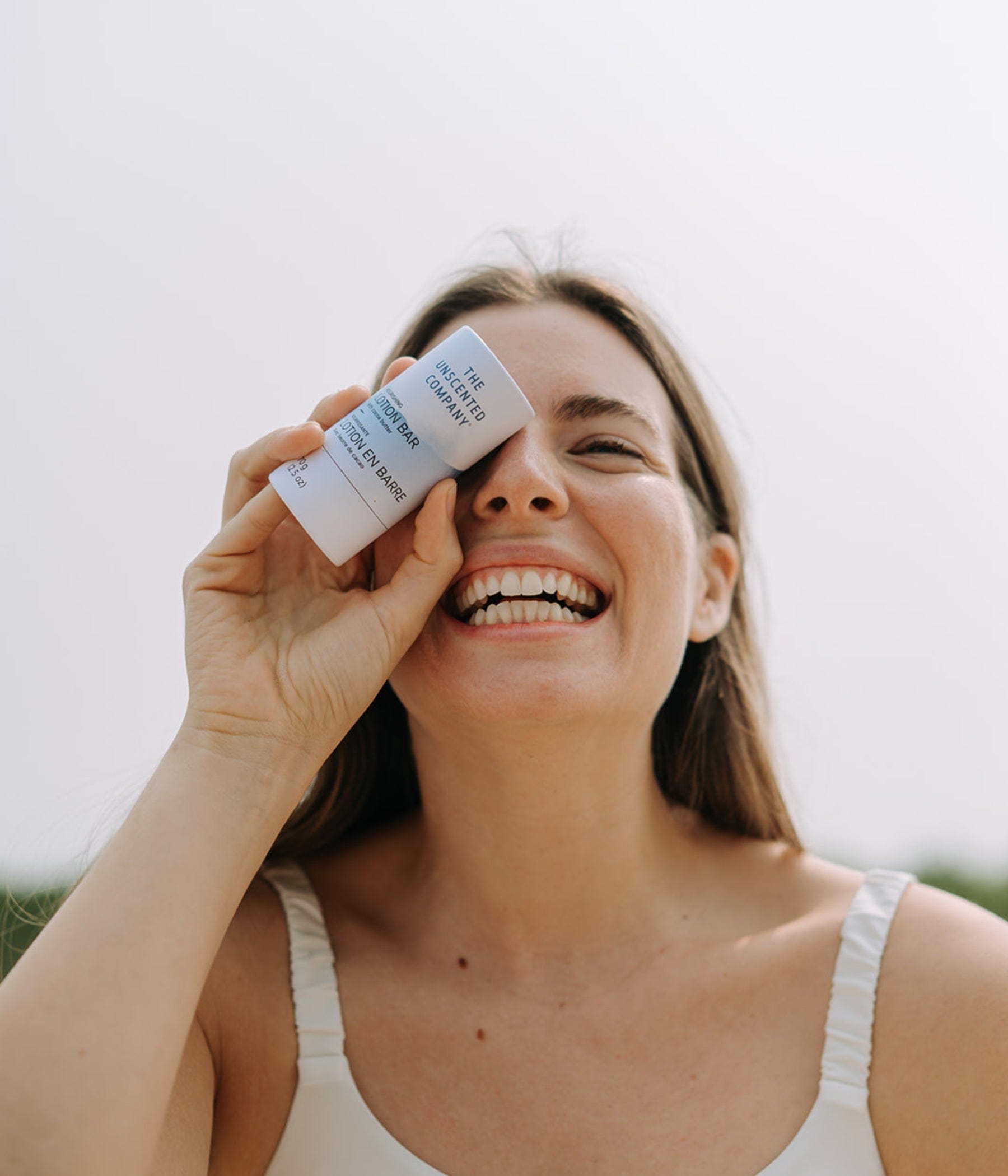 Smiling woman holding The Unscented Company’s lotion bar playfully in front of her eye on a sunny day.