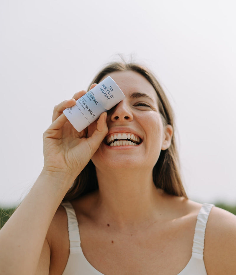 Smiling woman holding The Unscented Company’s lotion bar playfully in front of her eye on a sunny day.