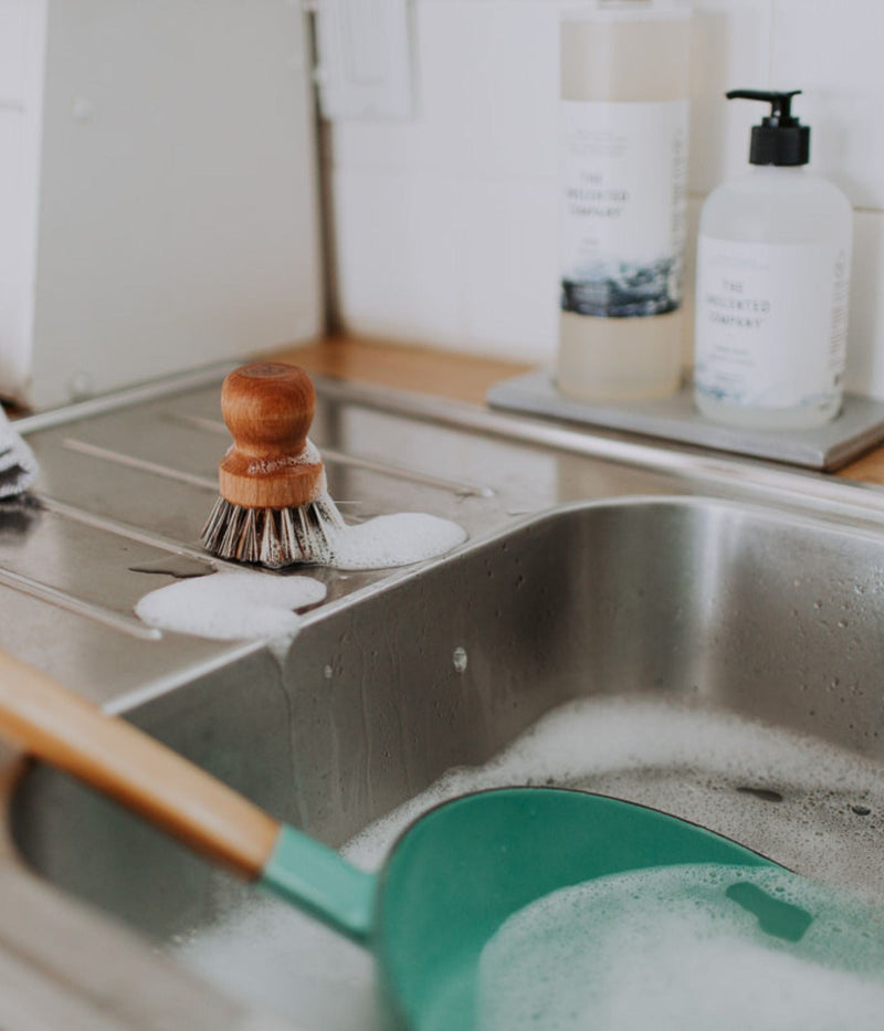 Wooden dish brush with soap suds by a sink, with The Unscented Company dish soap and hand soap in the background.