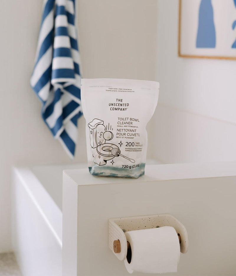 Un sachet de nettoyant pour cuvette de toilette The Unscented Company (200 pastilles) posé sur le rebord d’une baignoire blanche. En arrière-plan, une serviette rayée bleue et blanche est accrochée près d’un cadre au style minimaliste.