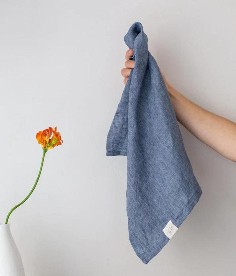Main tenant un linge à vaisselle en lin bleu à côté d’une fleur orange dans un vase blanc.