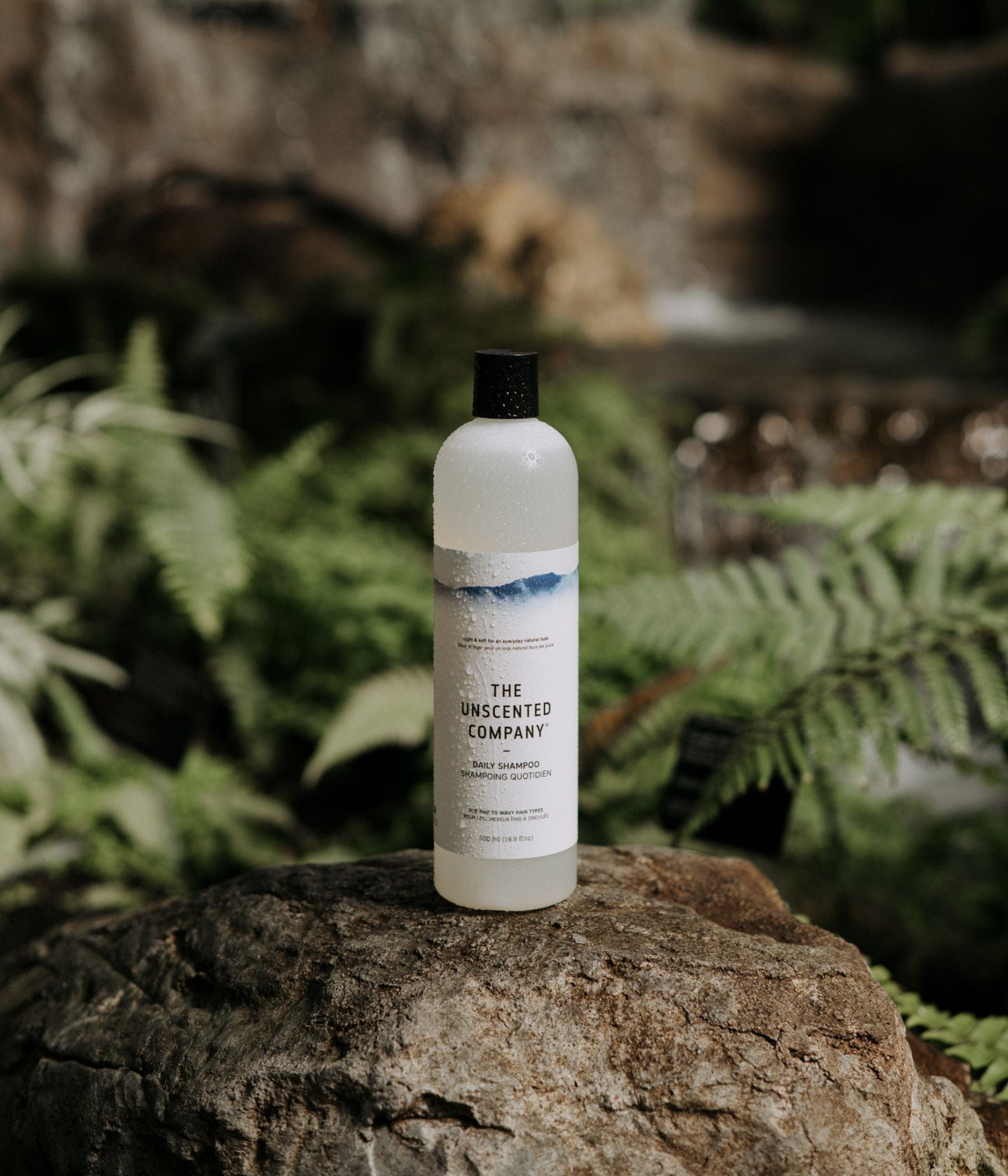 Shampoo bottle from The Unscented Company placed on a rock in a lush green setting.