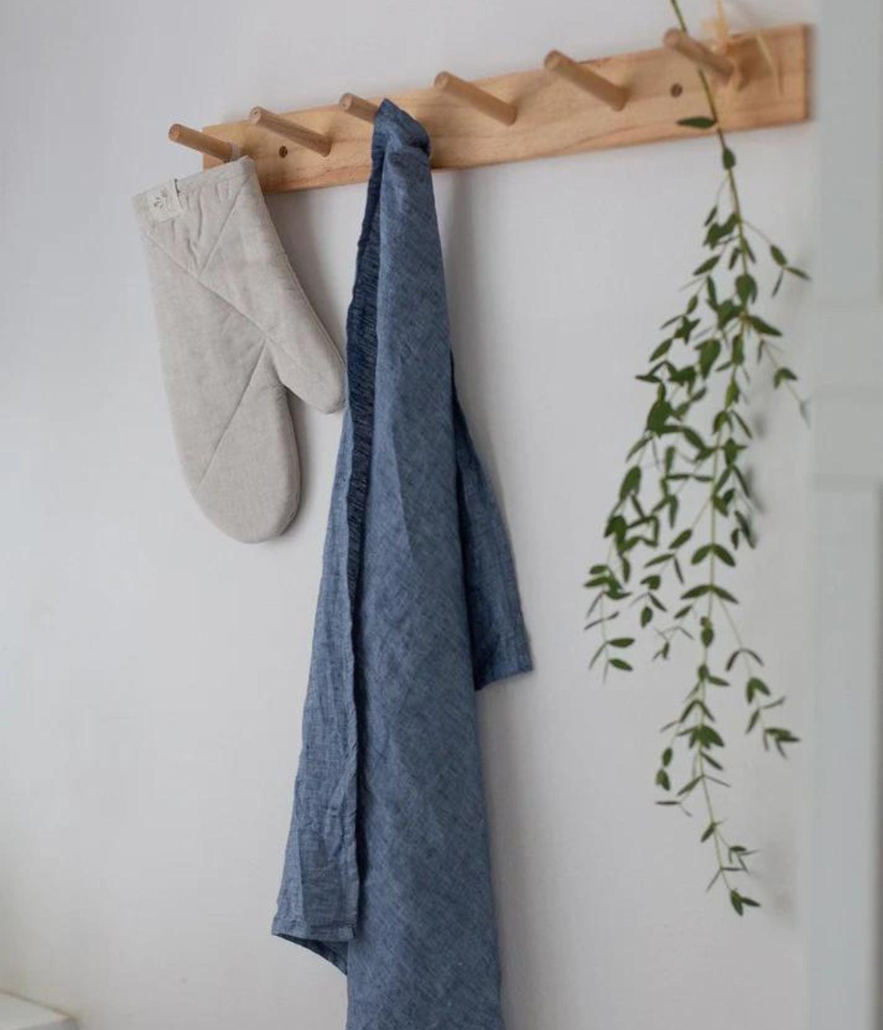 Blue linen dish towel and beige oven mitt hanging on a wooden peg rack with a green leafy branch nearby.