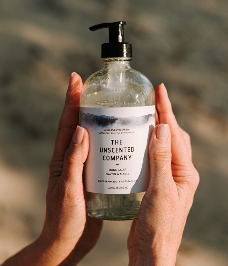 A bottle of The Unscented Company hand soap held with both hands in soft light.