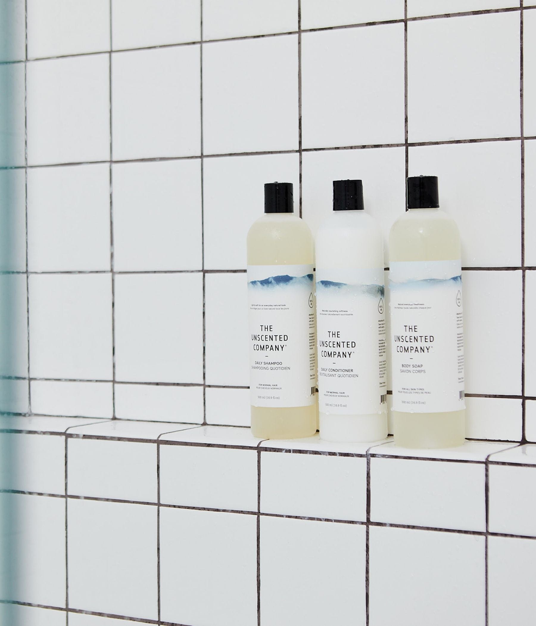 Trois bouteilles de produits The Unscented Company—shampoing, revitalisant et savon pour le corps—alignées sur un rebord de douche carrelé.