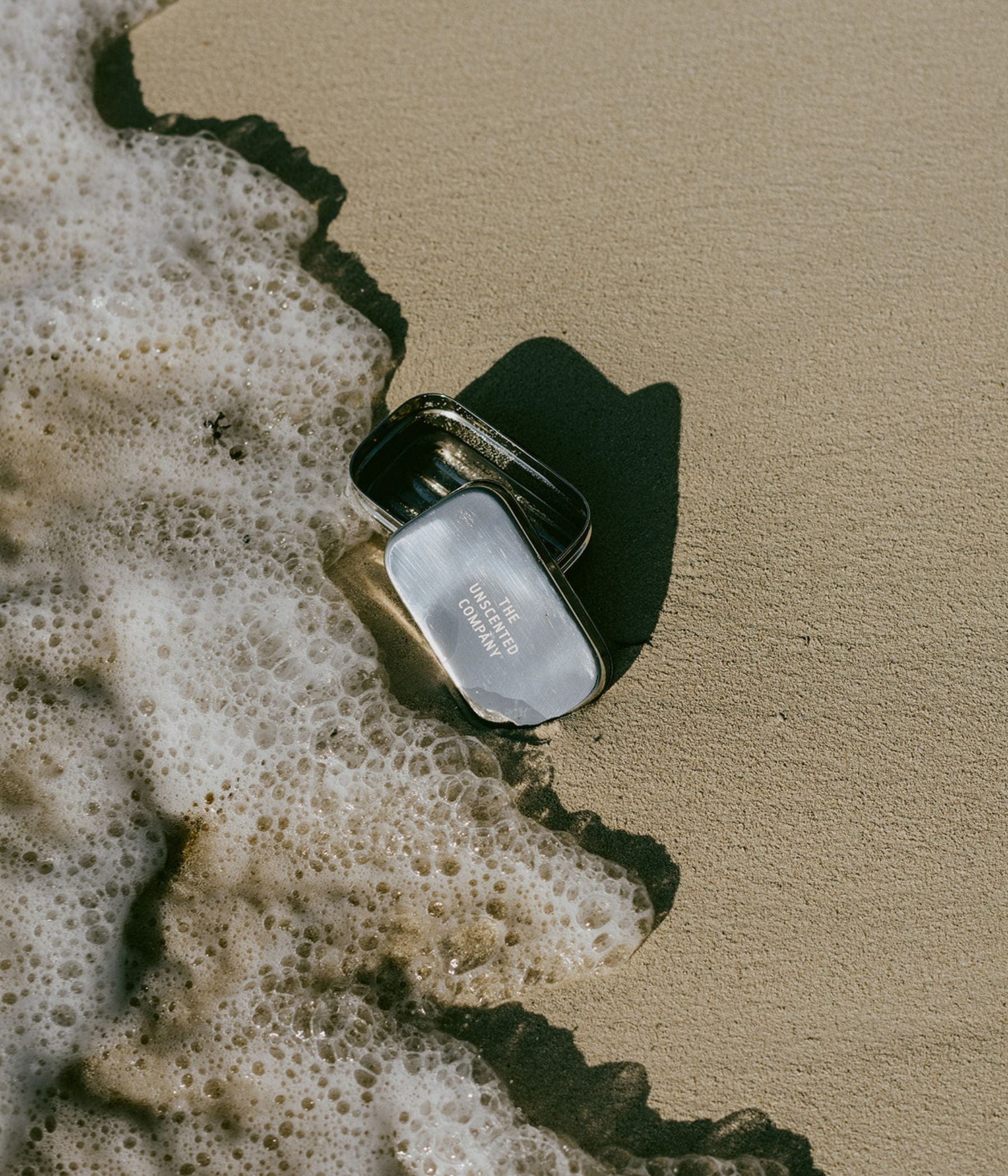Open metal tin from The Unscented Company lying on wet sand, with sea foam gently reaching it.