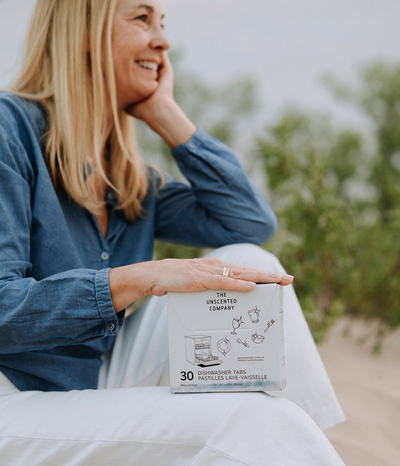 Une femme assise sur le sable sourit tout en tenant une boîte de pastilles lave-vaisselle The Unscented Company.