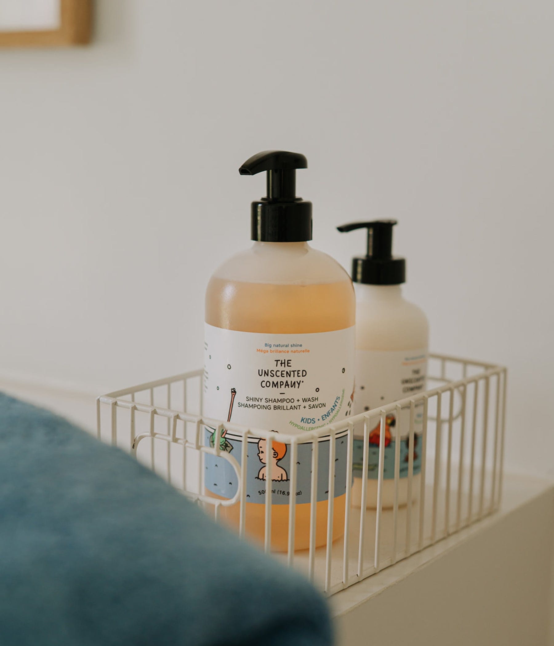 The Unscented Company Shiny Shampoo + Wash for kids in a reusable pump bottle, displayed in a white wire basket in a bright bathroom.