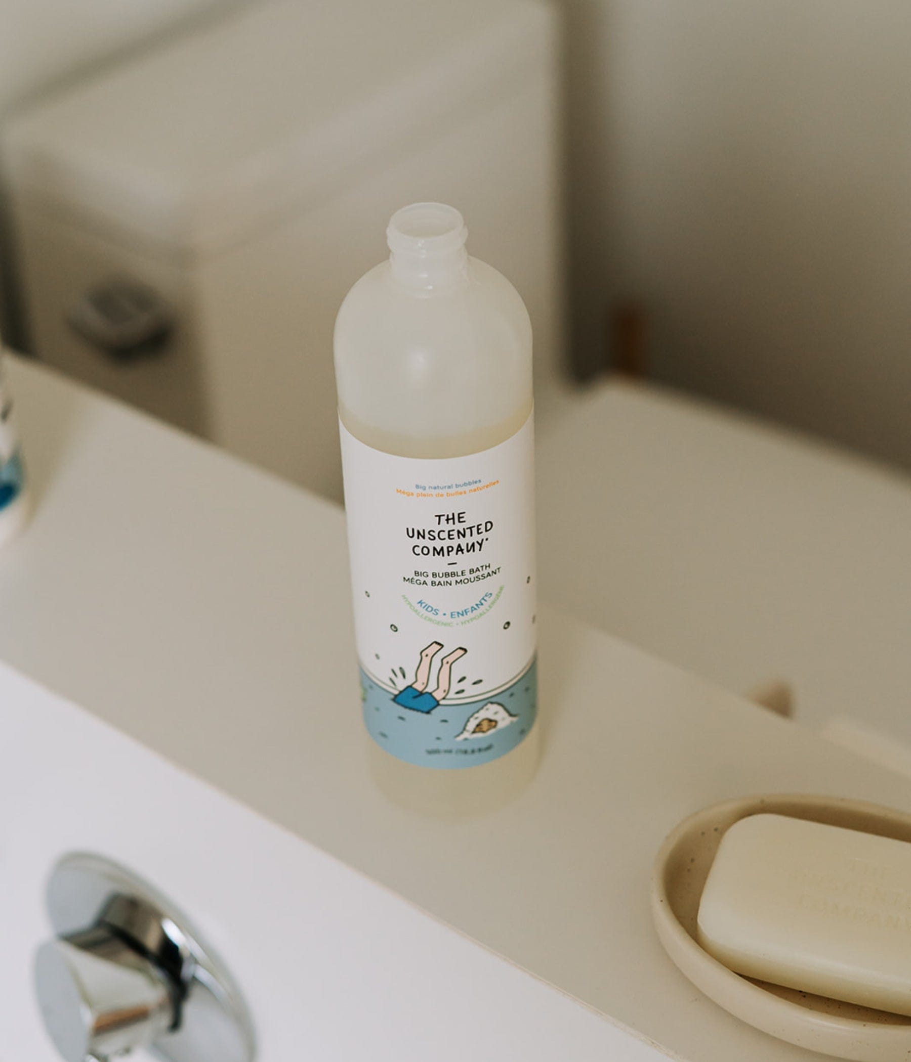 The Unscented Company Big Bubble Bath for kids in a clear bottle, placed on a bathtub ledge beside a natural soap bar in a minimalist bathroom.