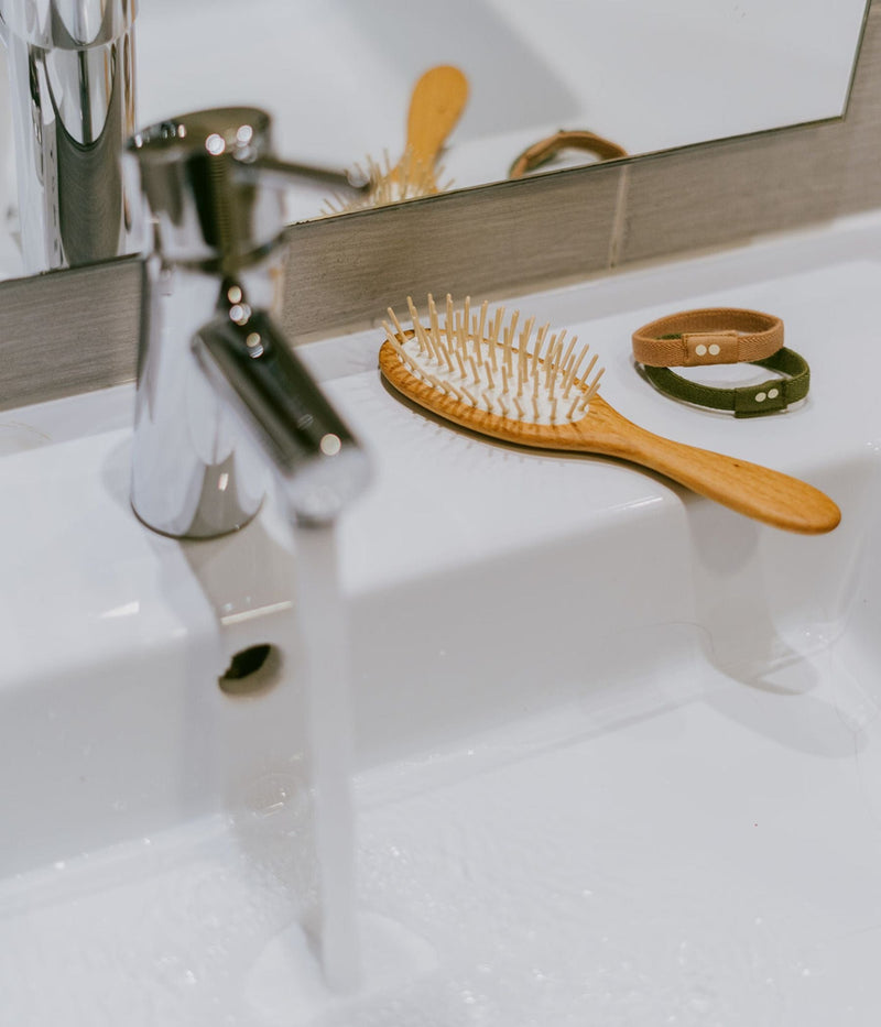 Wooden hairbrush and fabric bracelets on a bathroom sink with running water.