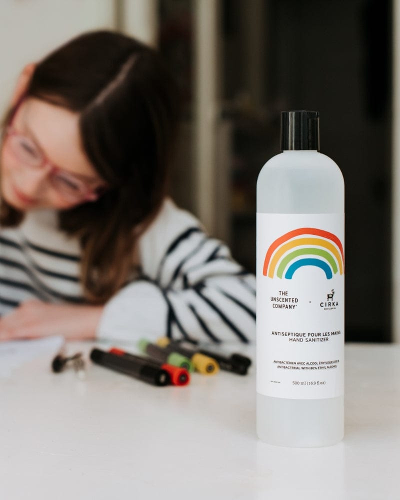 the Unscented Company hand sanitizer bottle on a table with a child drawing in the background.