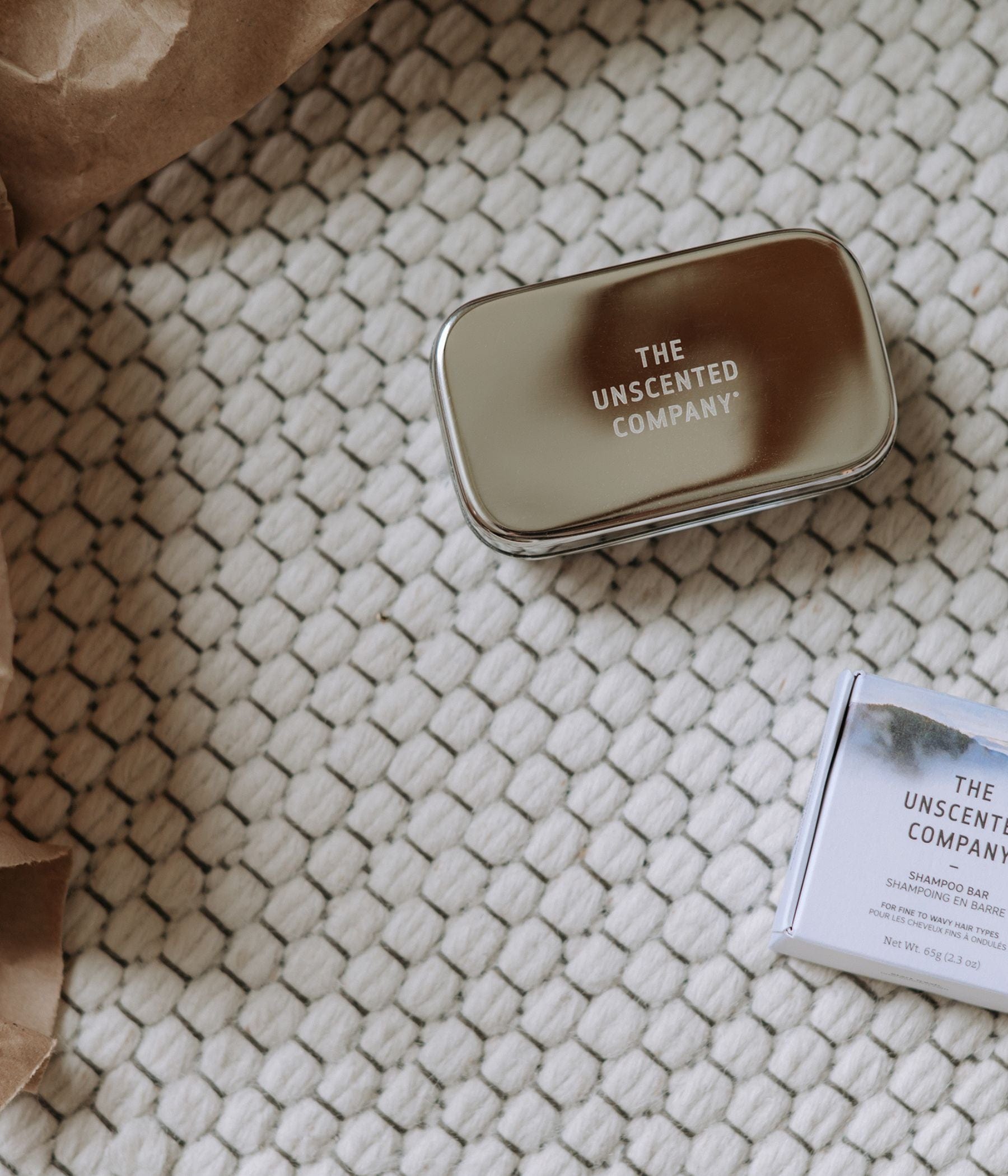 Closed silver tin from The Unscented Company on a woven cream rug, next to a boxed shampoo bar.