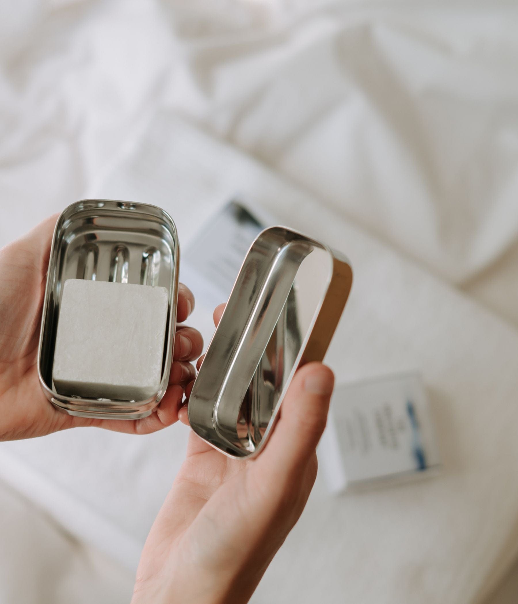Hands holding an open metal tin with a white soap block inside, placed on soft white fabric.