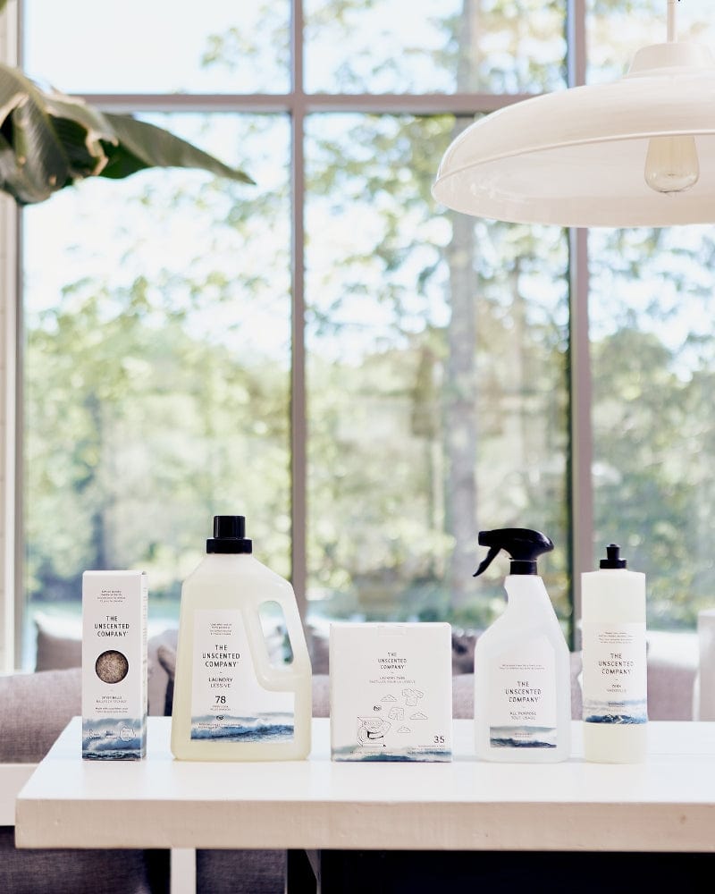 The Unscented Company products, including laundry detergent, dryer balls, cleaning spray, and tabs, arranged on a white table in front of a bright window.