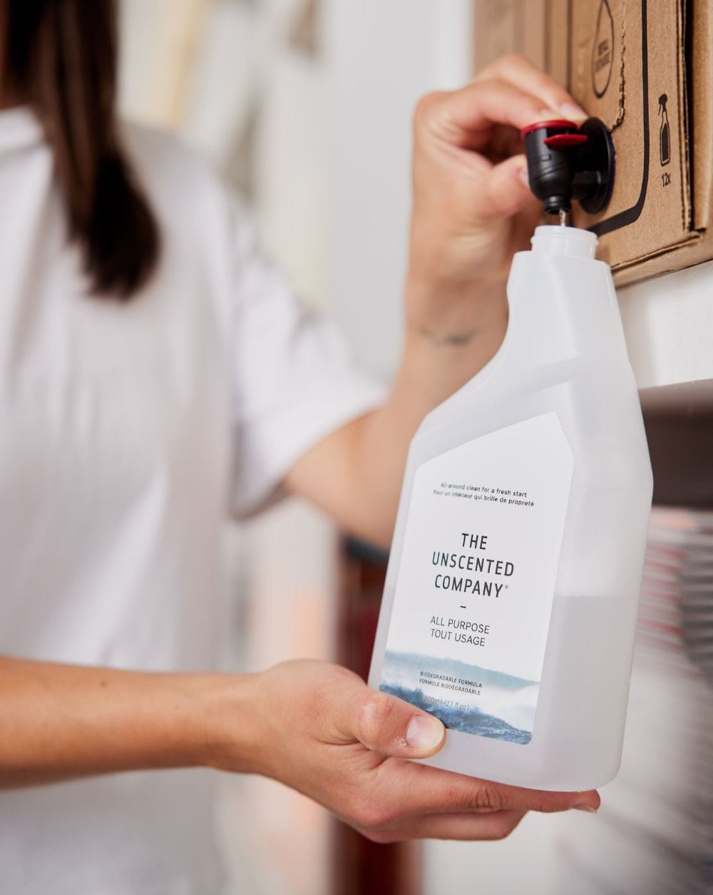 Person refilling a The Unscented Company all-purpose cleaner bottle from a cardboard dispenser.