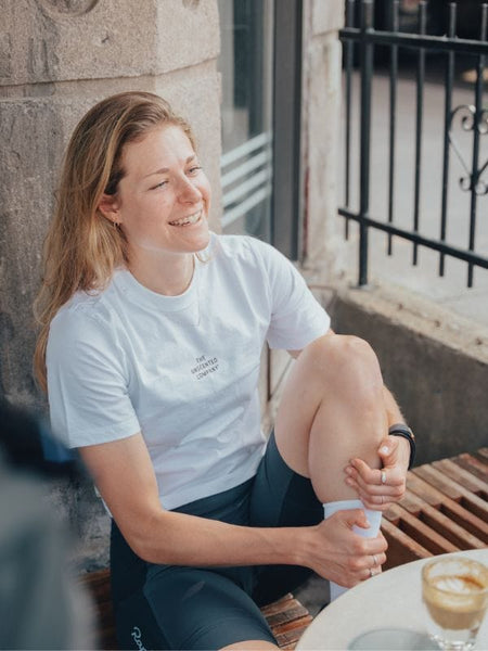 Woman wearing a white The Unscented Company t-shirt smiling while sitting at an outdoor café.