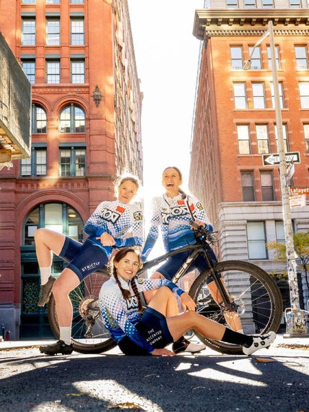 Three women in cycling kits posing with a bicycle on a city street.