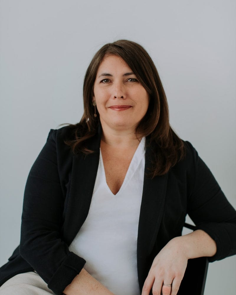 Woman in a white shirt and black blazer sitting and smiling in front of a plain background.
