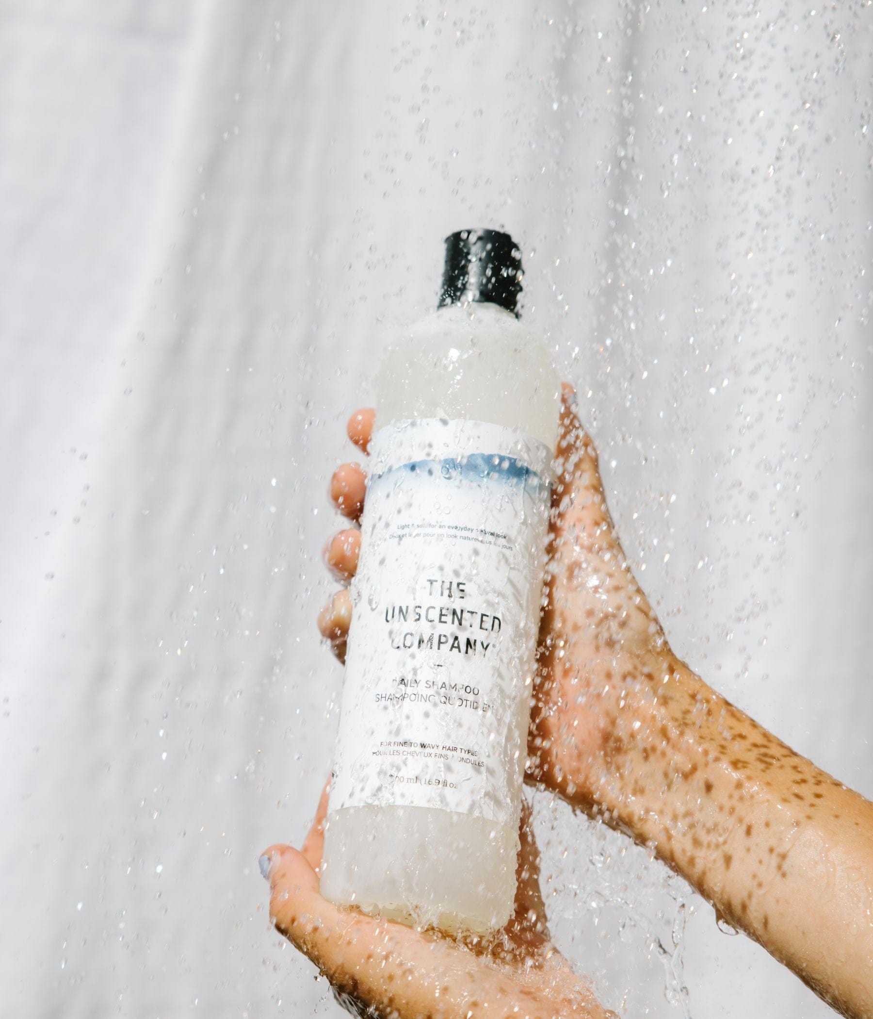 Hands holding a bottle of The Unscented Company shampoo under running water