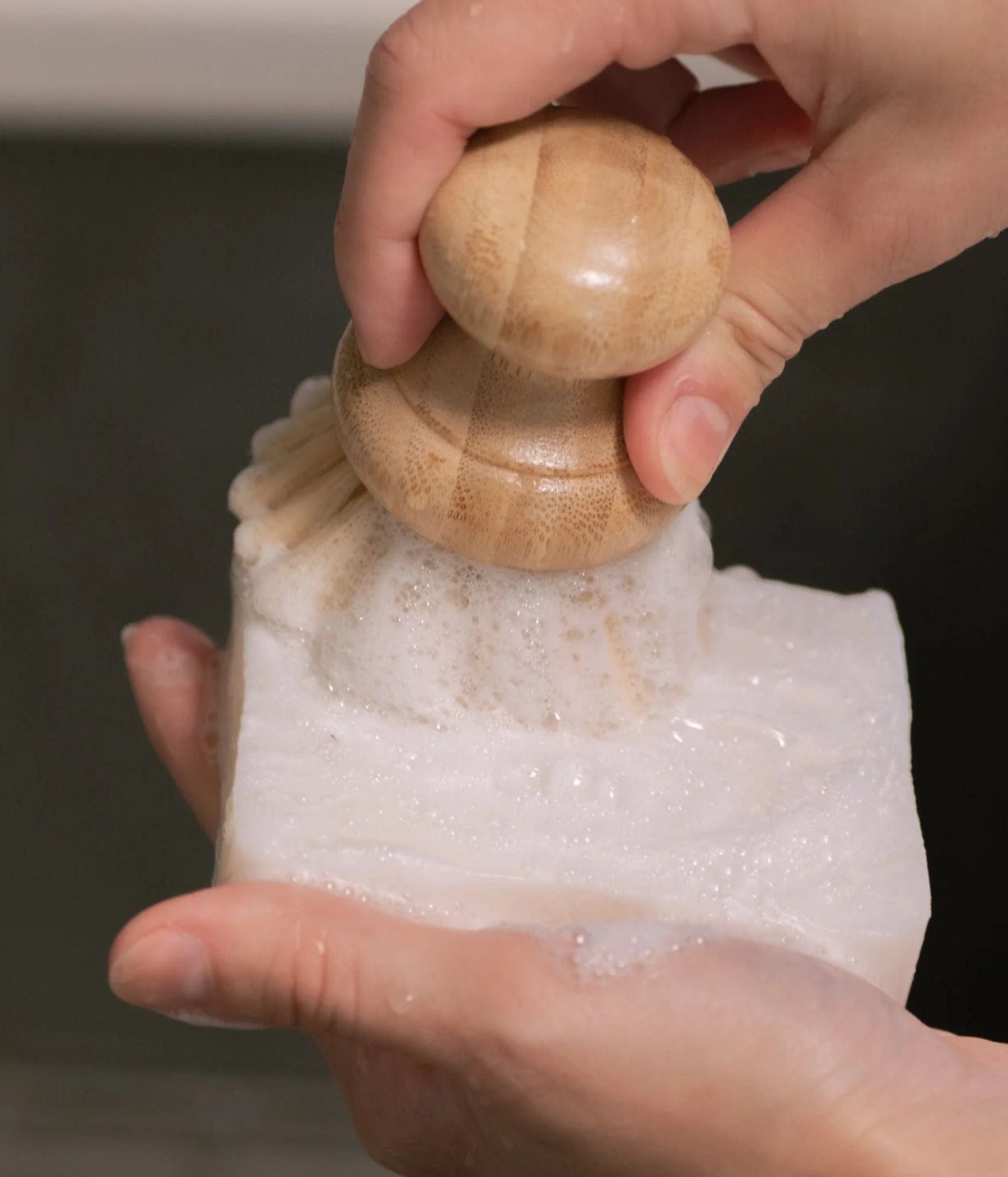 Hand brushing a dish soap block with a round bamboo brush.