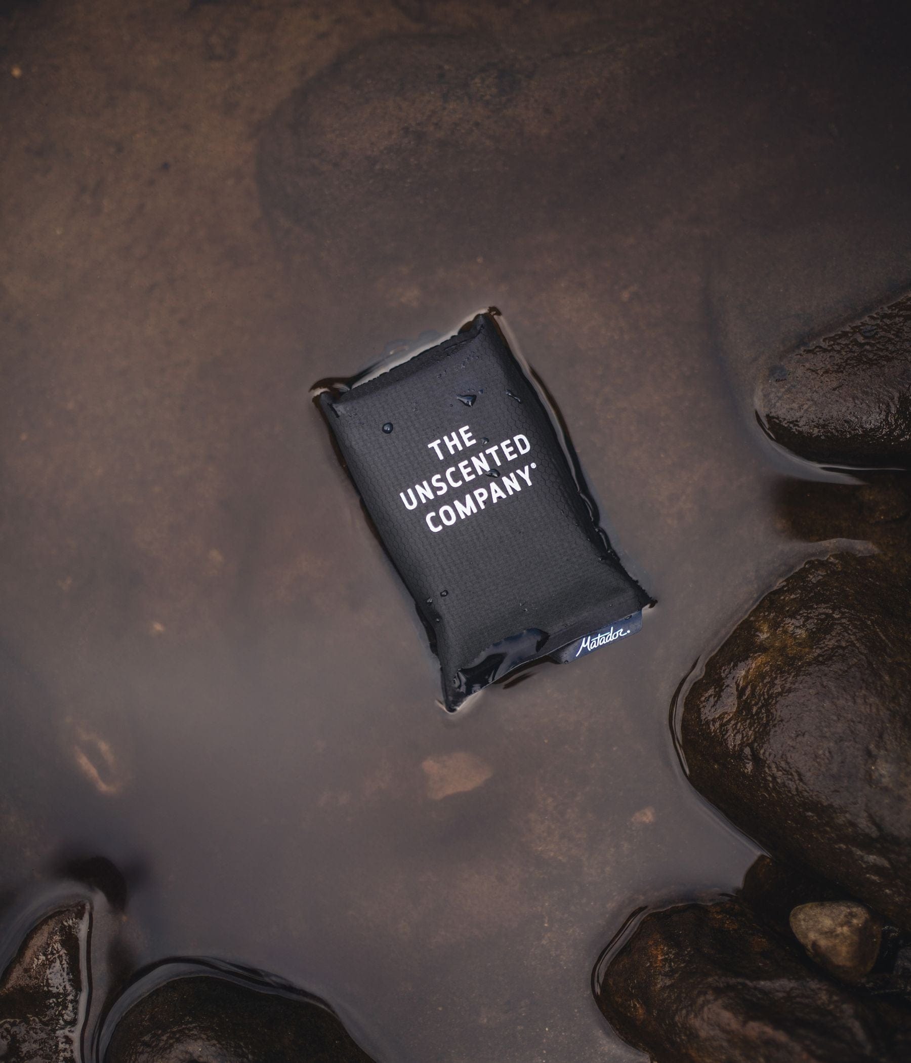 The Unscented Company pouch floating in water among rocks.