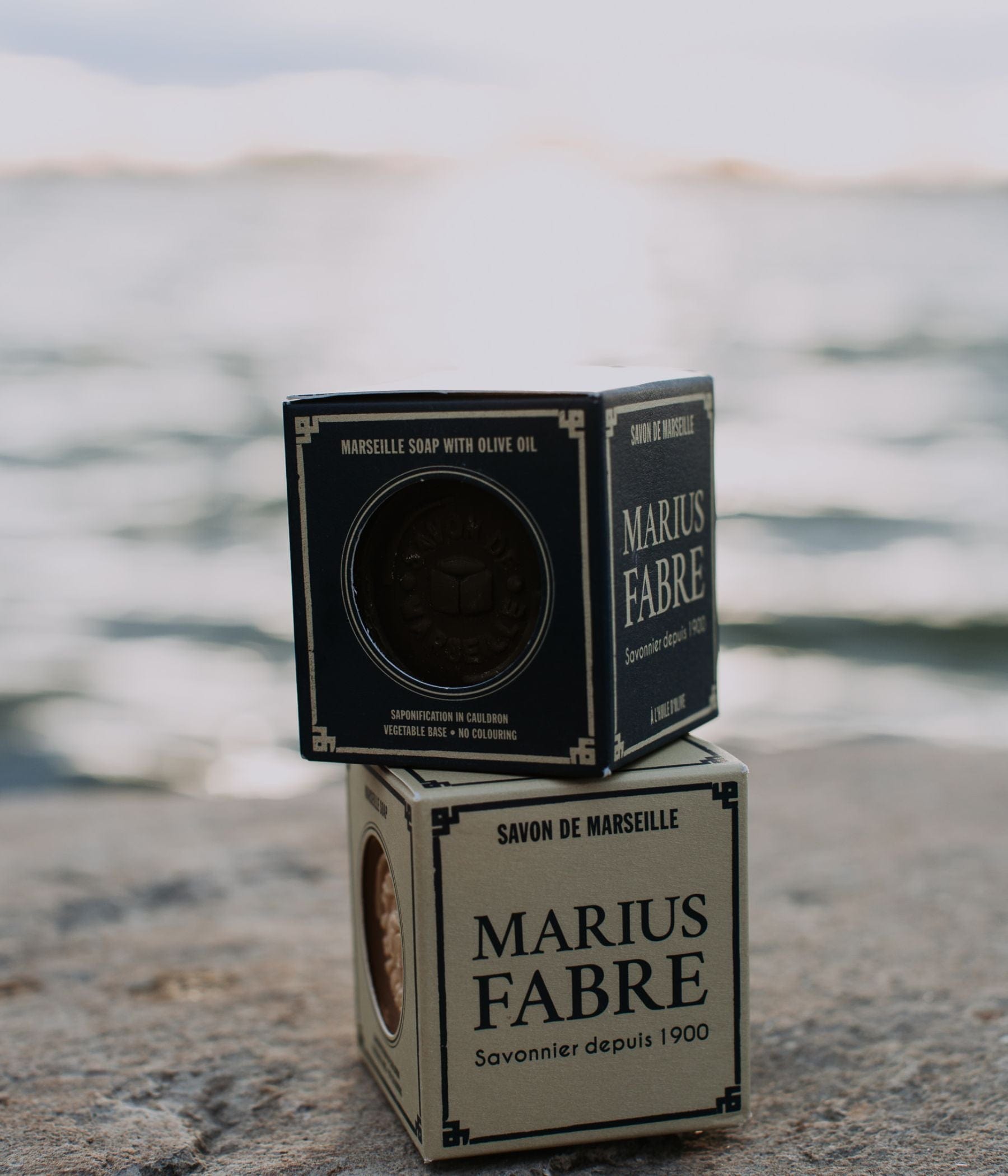 Two boxes of Marius Fabre Marseille soap stacked on a rock by the water.
