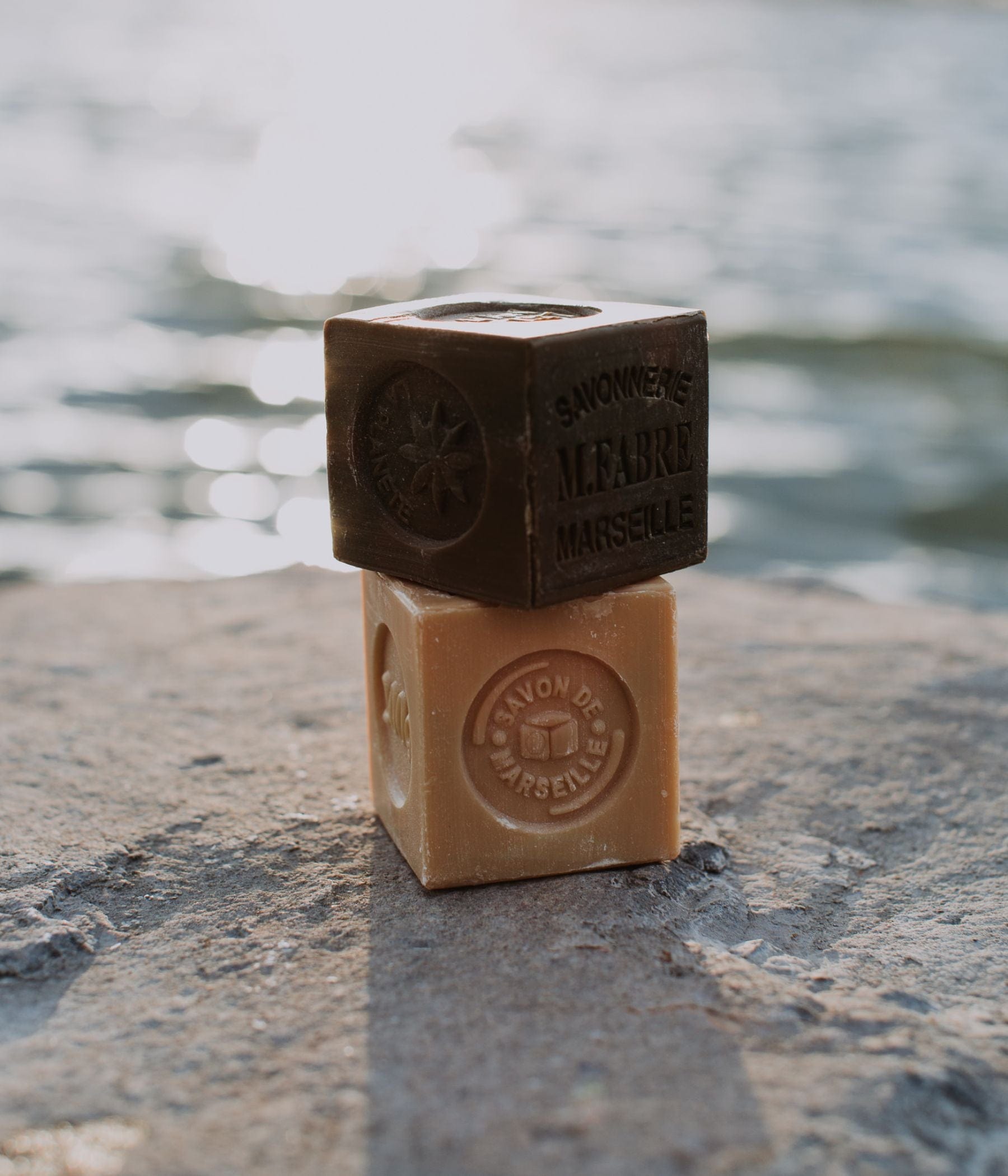 Two Marseille soap cubes stacked on a rock by the water.