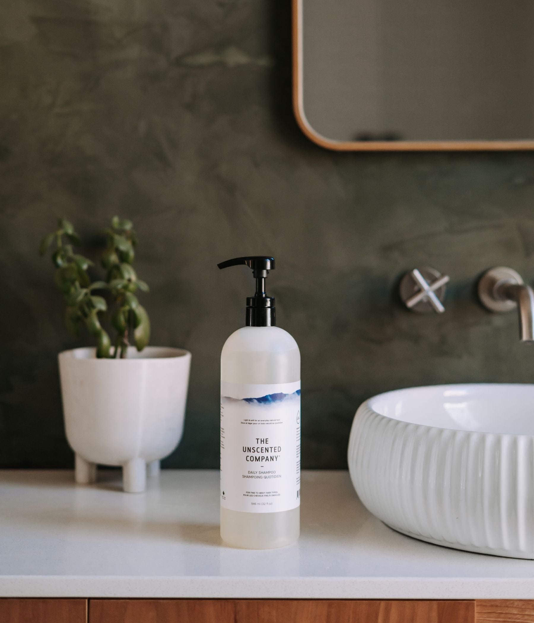 The Daily Shampoo - 1L placed a bathroom counter, on the left side of a sink, dark green background.