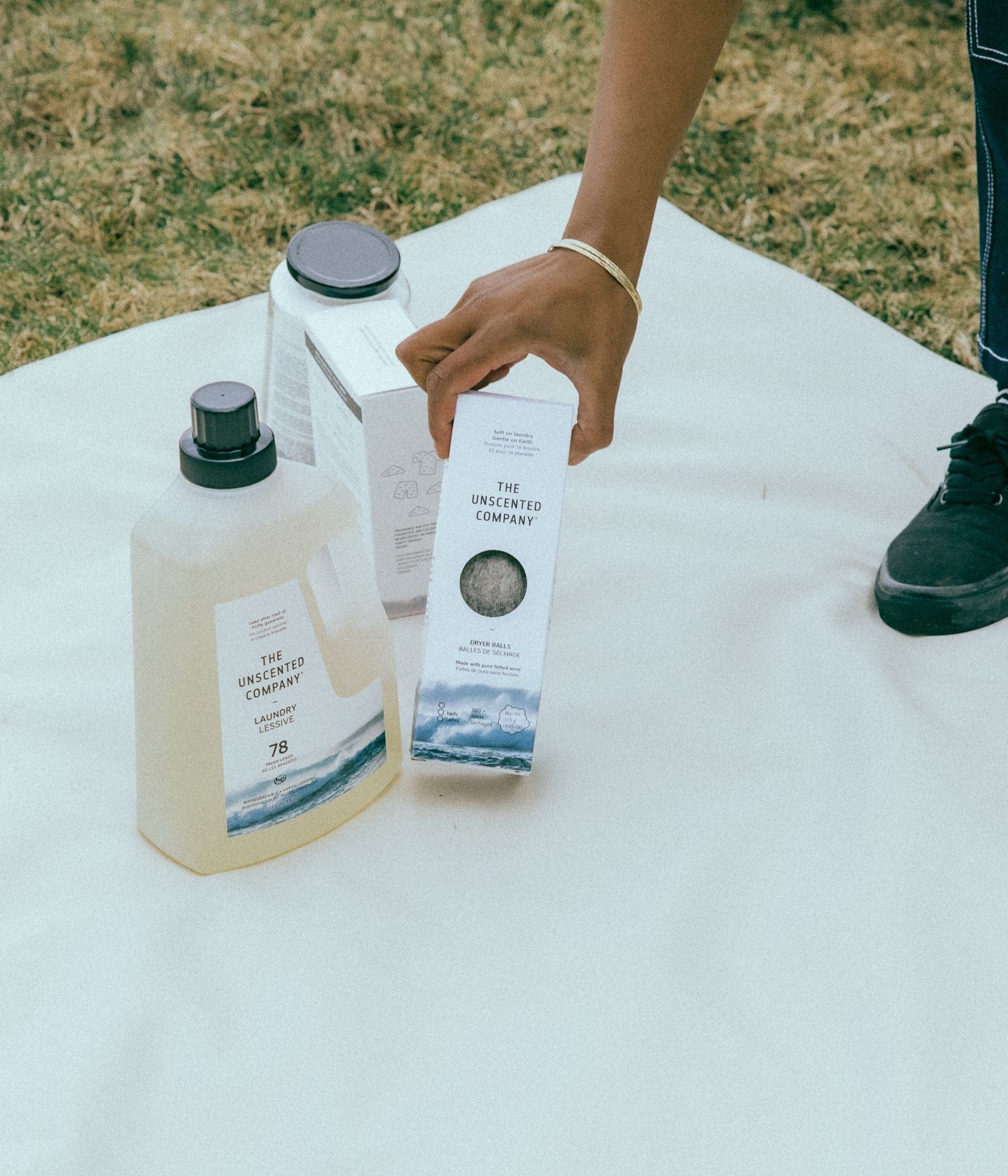 Person picking up a box of dryer balls next to laundry detergent and tabs on a white cloth outdoors.
