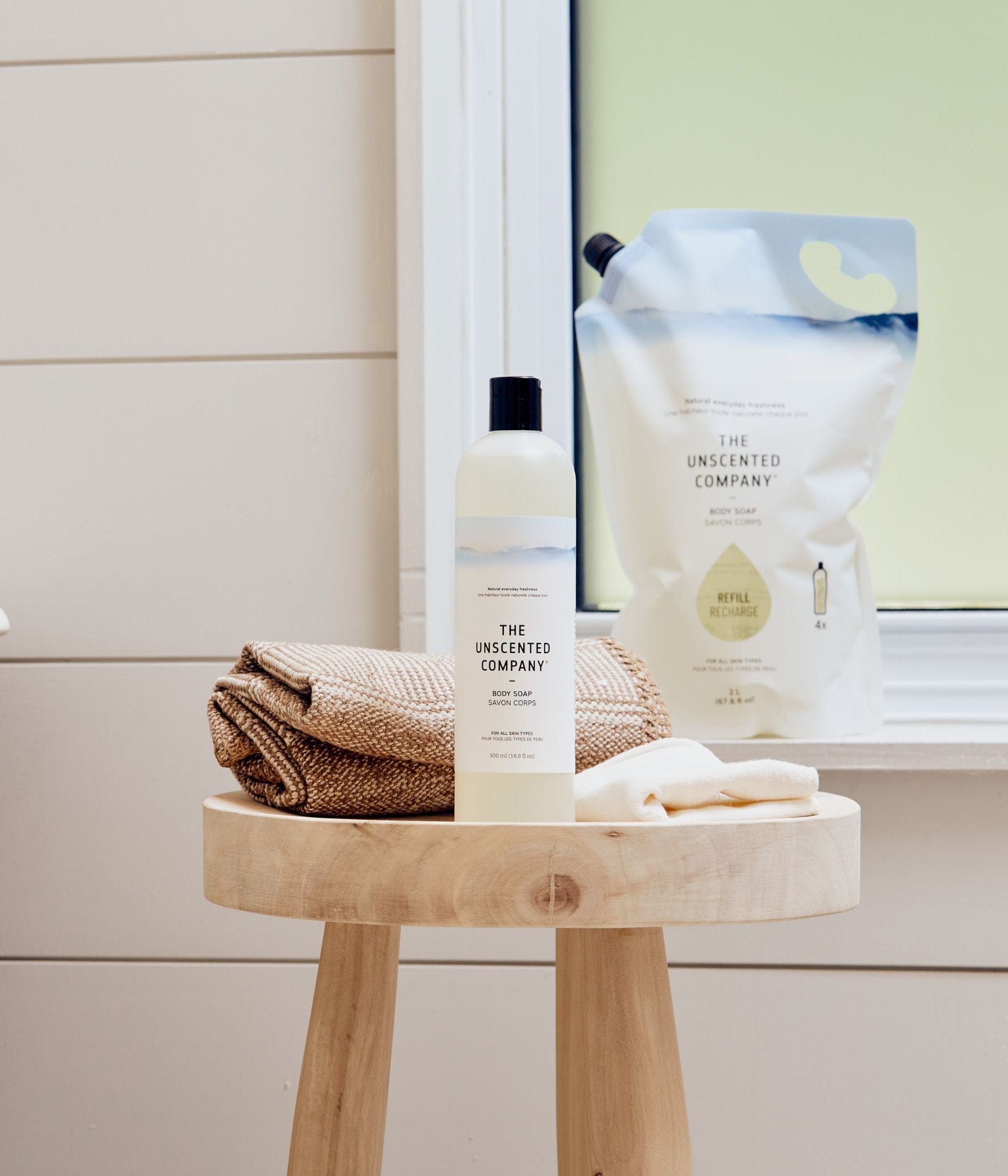 Body soap bottle and refill pouch from The Unscented Company on a bathroom stool.