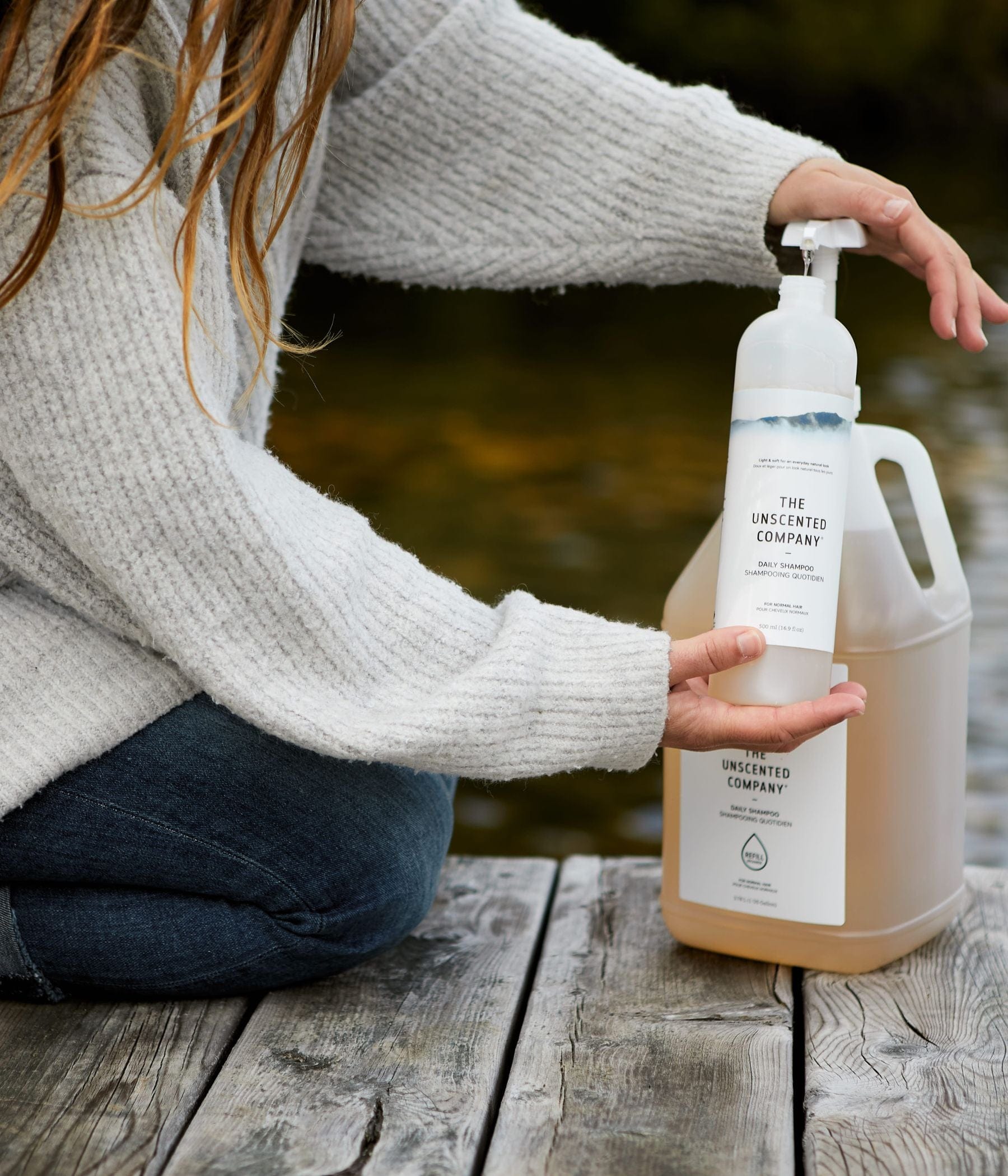 Person filling a shampoo bottle from a large The Unscented Company container on a dock.