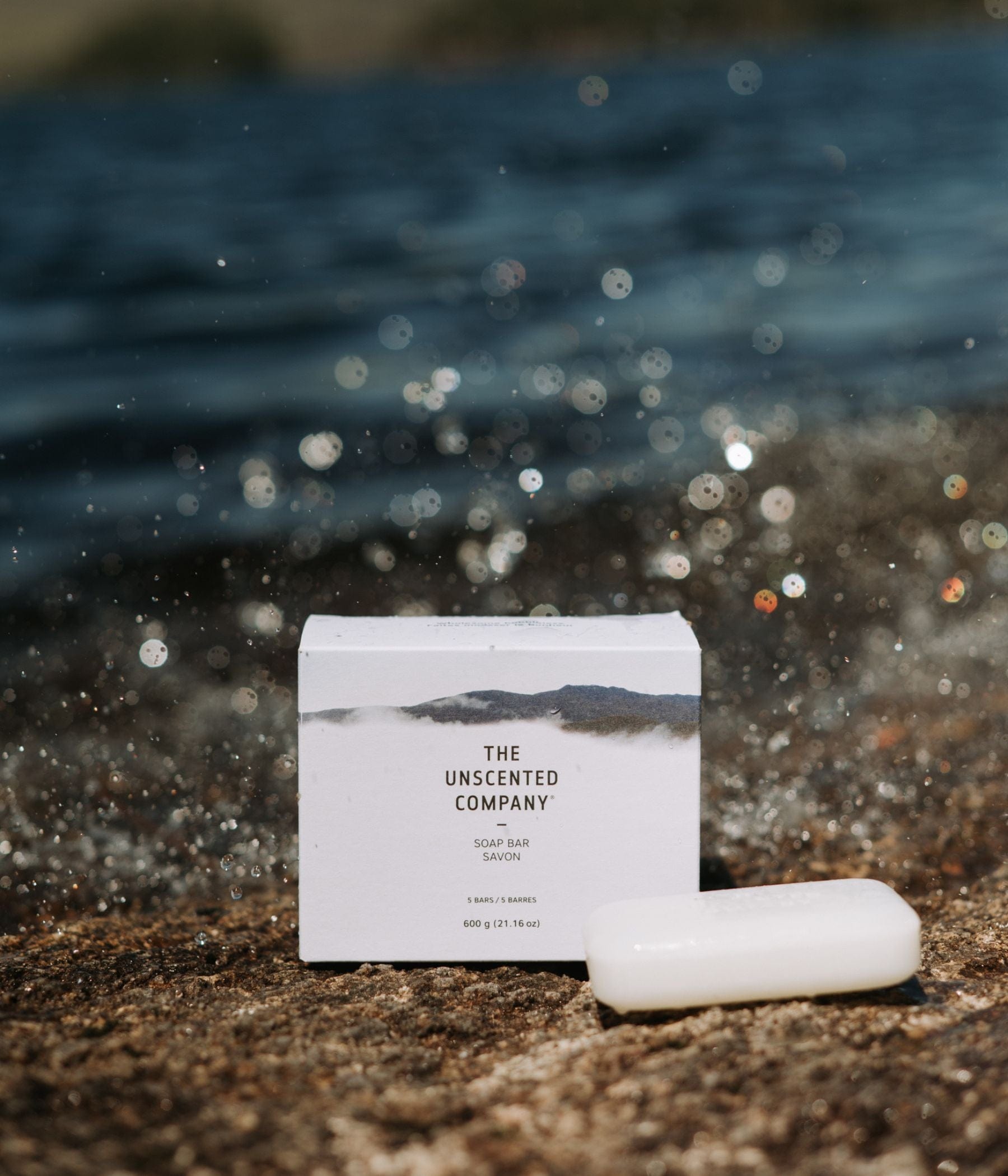 The Unscented Company soap bar and box on a rock near water.