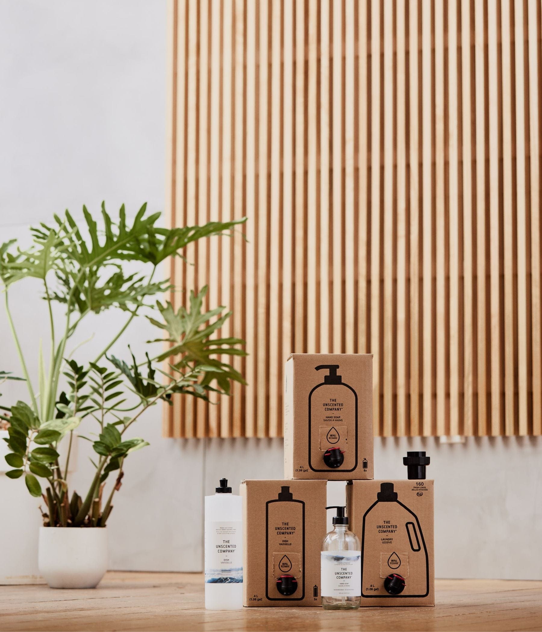 Cleaning products from The Unscented Company stacked on a wooden floor with a plant in the background.