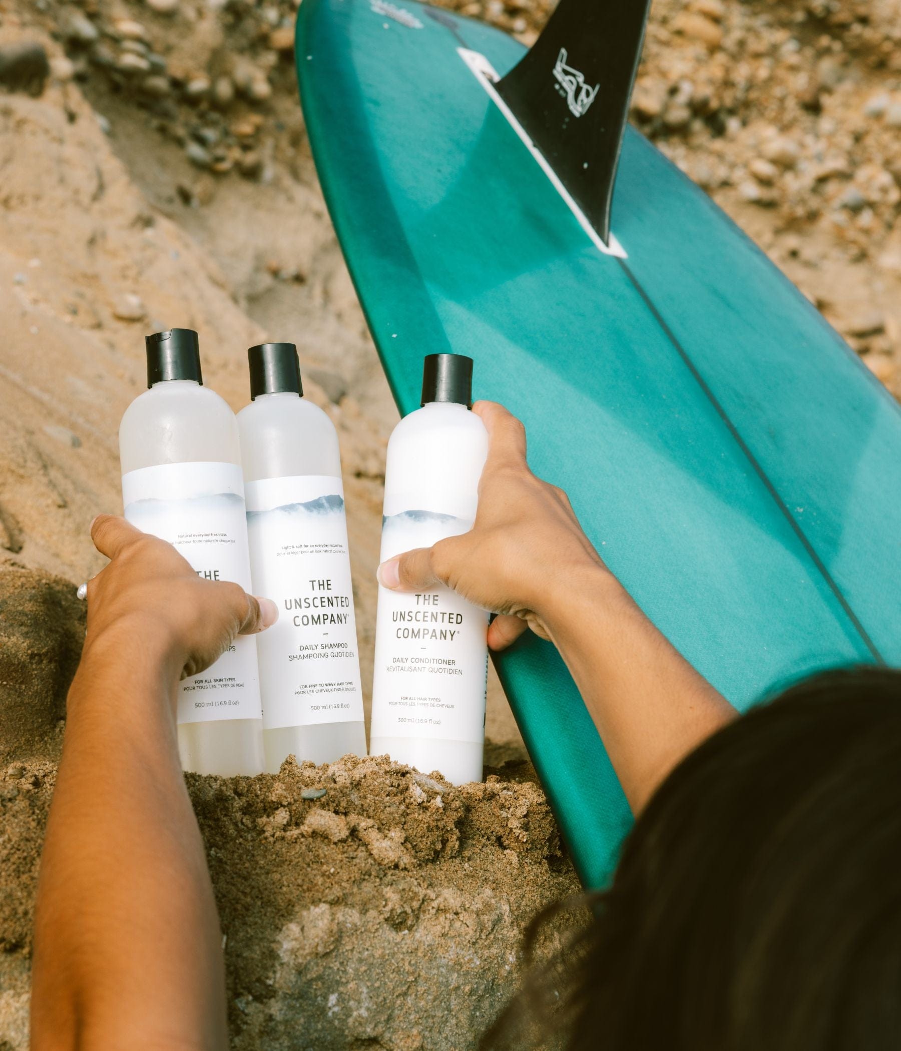 Une personne tient trois bouteilles de shampooing et revitalisant The Unscented Company dans le sable à côté d’une planche de surf turquoise.