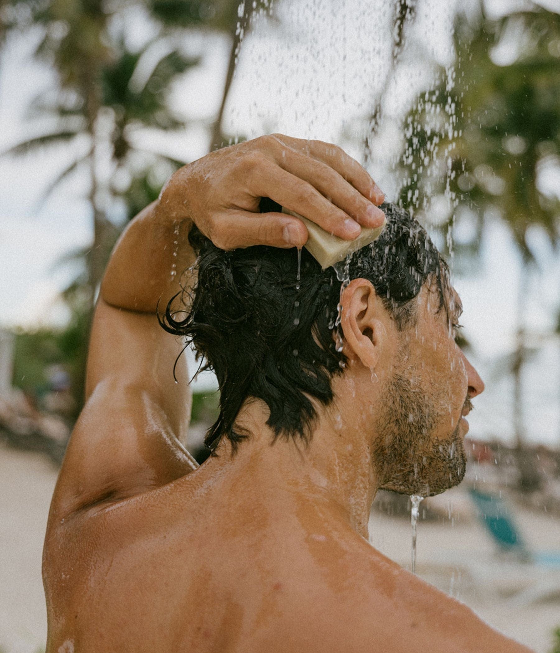 A man outdoors uses a bar of soap to wash his hair under running water, with palm trees in the background.