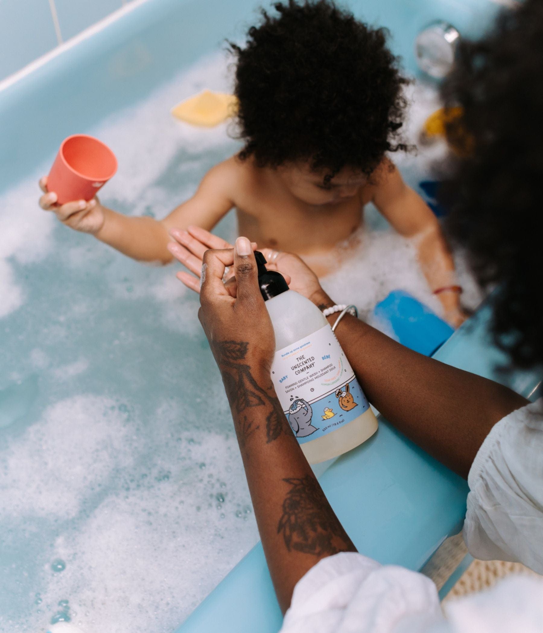 Adult dispensing baby body wash into hand while child plays in a bubble bath.