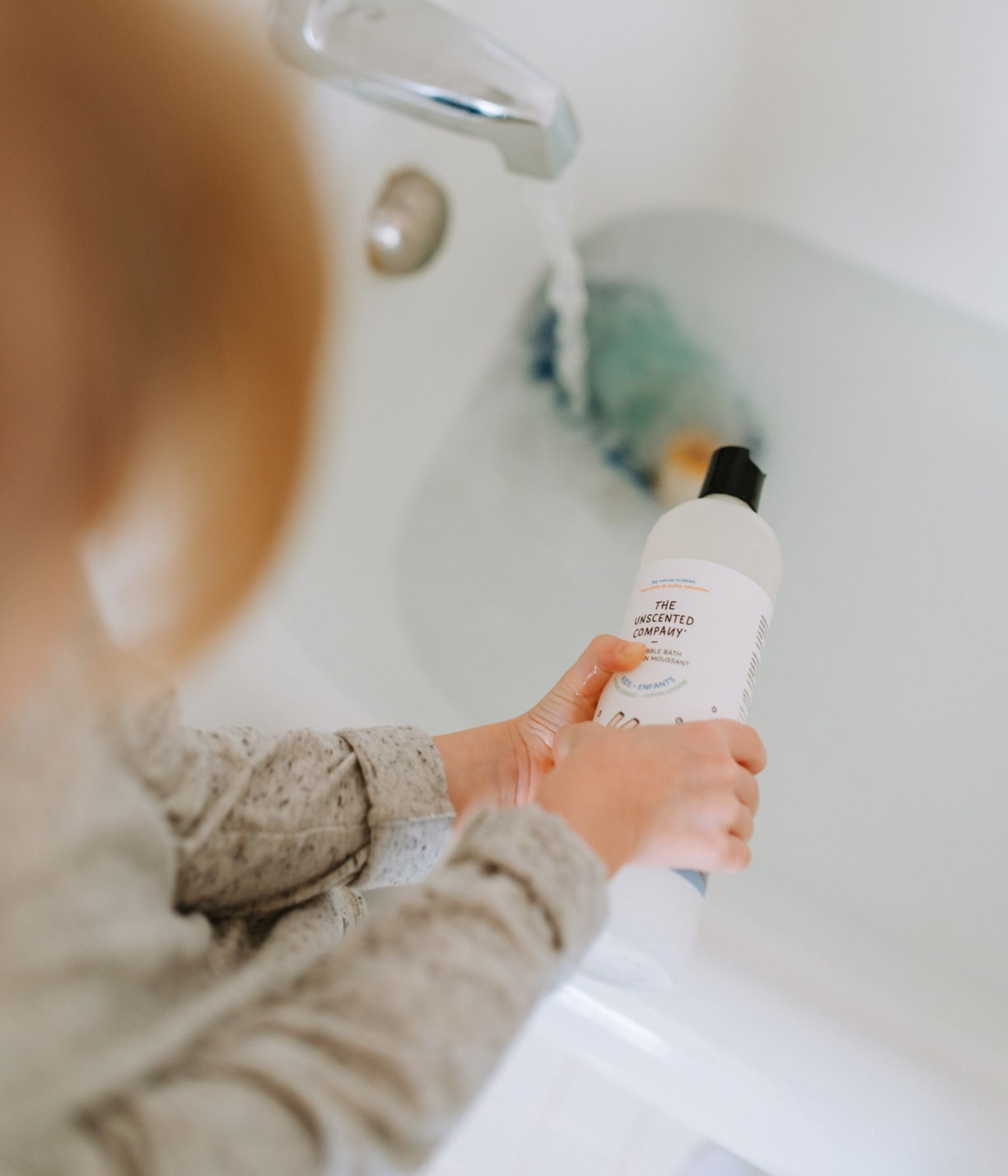 Child holding a bottle of The Unscented Company next to a running bath.