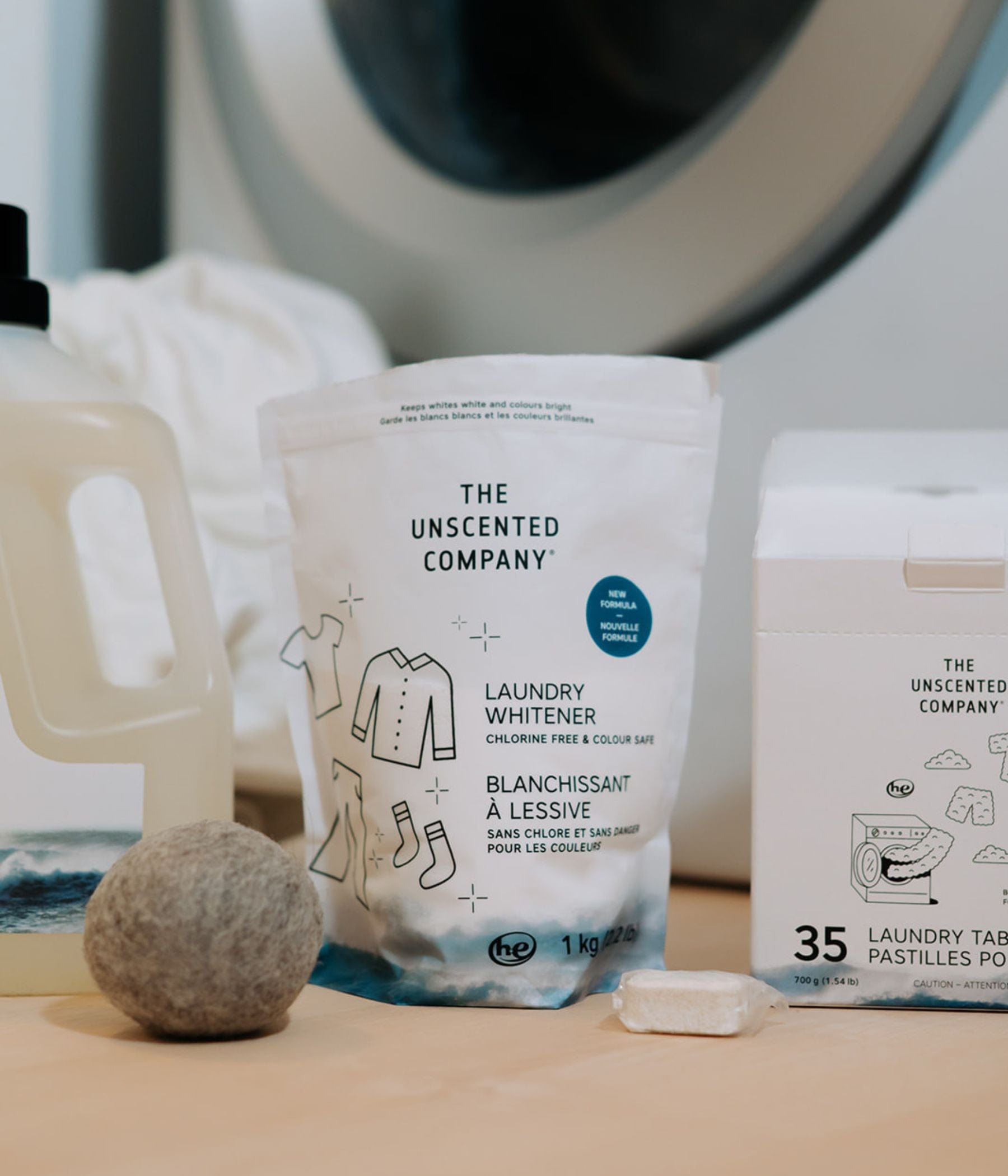 The Unscented Company’s laundry care products in front of a washing machine: a bottle of detergent, a bag of laundry whitener, and a box of 35 laundry tabs. A dryer ball and an unwrapped laundry tab are placed in front.