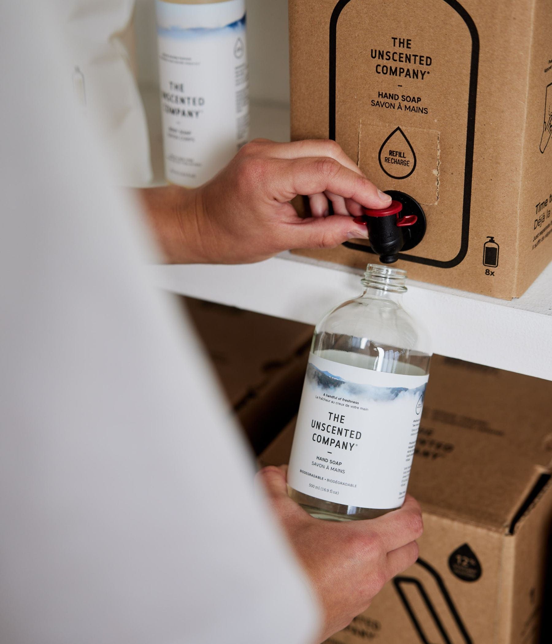 A person refilling a The Unscented Company hand soap bottle from a cardboard dispenser.