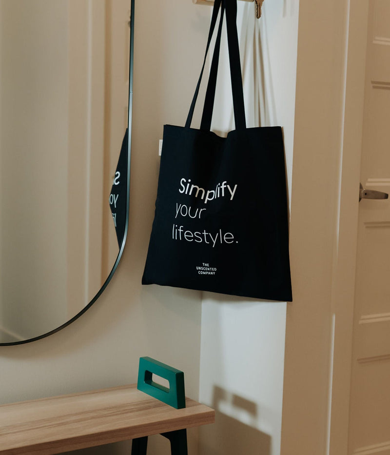 A black tote bag hanging on a wall hook in a hallway. The bag has white text that reads “Simplify your lifestyle.” with The Unscented Company logo below.