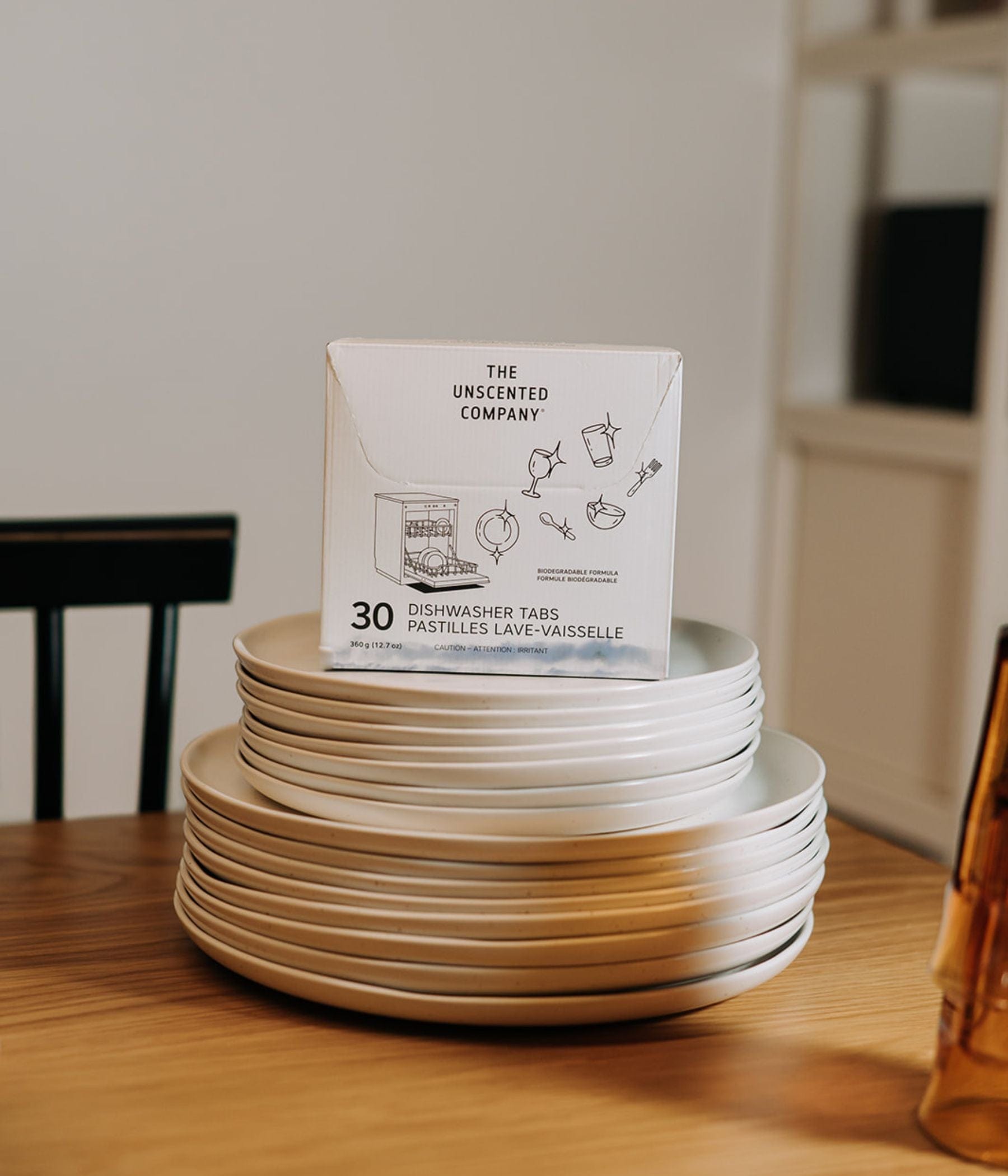 Box of 30 The Unscented Company dishwasher tabs placed on a stack of white plates on a wooden table.