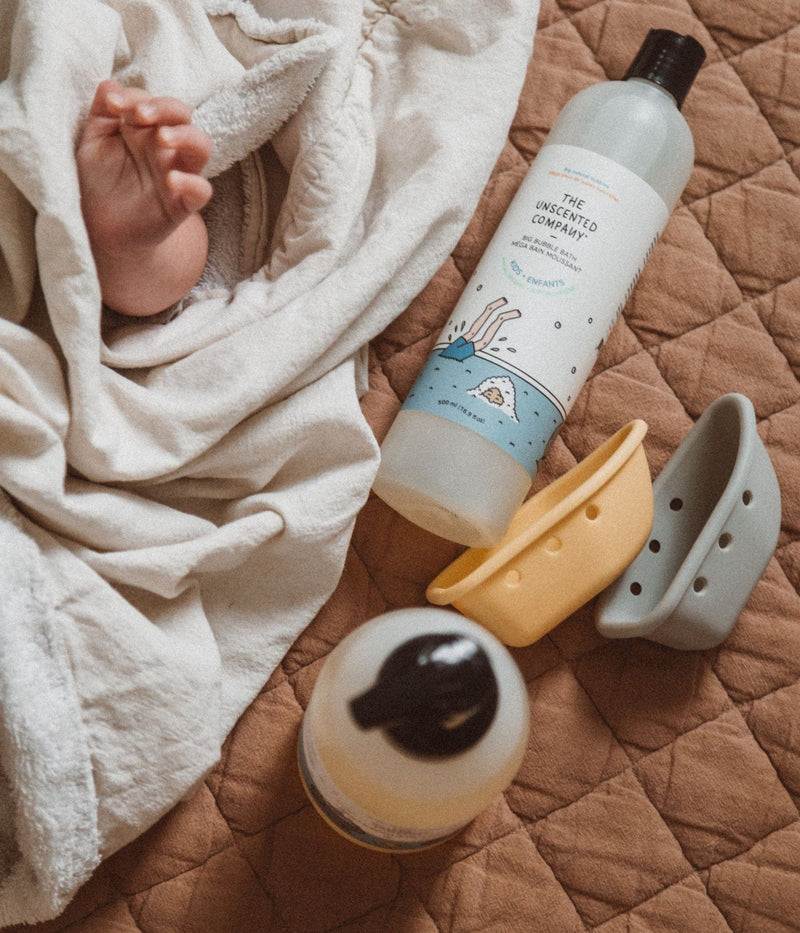 Baby wrapped in a blanket with The Unscented Company baby products and toy boats.