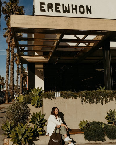 A woman wearing sunglasses and a white shirt sits on a wooden bench outside Erewhon Market, surrounded by lush greenery and palm trees under a sunny sky.