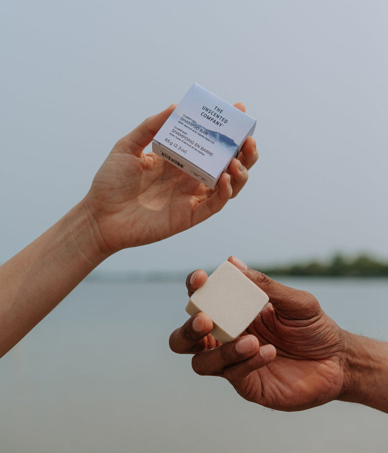 Two hands holding The Unscented Company’s shampoo bar and its box packaging, outdoors near a calm lake.