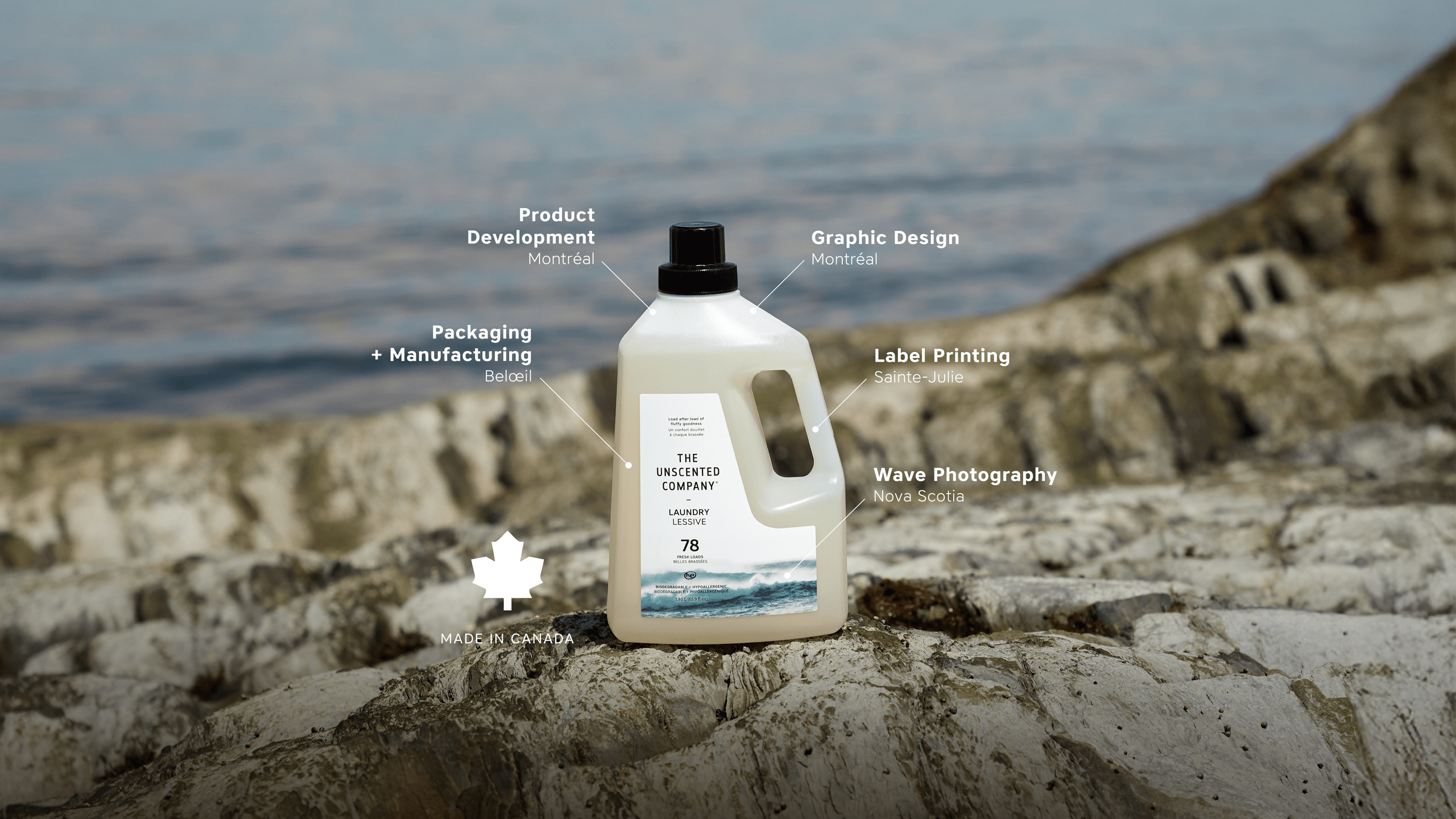 Bottle of The Unscented Company laundry detergent placed on a rocky shoreline with water in the background. Labels identify each production step: product development and graphic design in Montréal, packaging and manufacturing in Beloeil, label printing in Sainte-Julie, and wave photo used on the label taken in Nova Scotia. A “Made in Canada” badge with a maple leaf is shown at the bottom left.